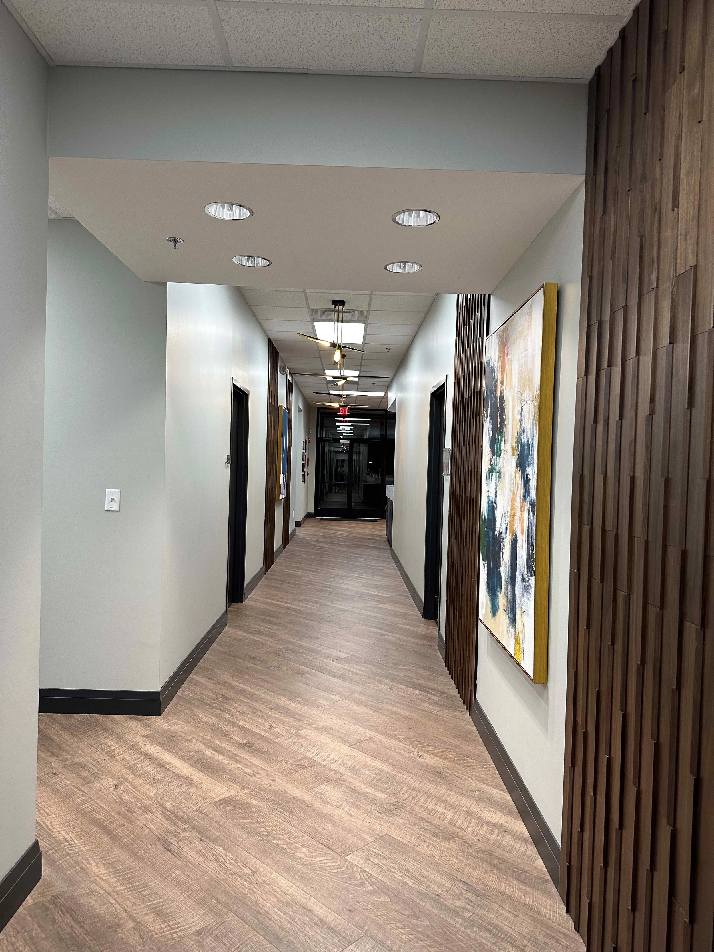 Corporate office secondary hallway with walnut wood slat accent wall, large abstract artwork in gold frame, brass angular pendant lights, wood plank floors, and dark framed office doors, interior design by Luxe Style Studio