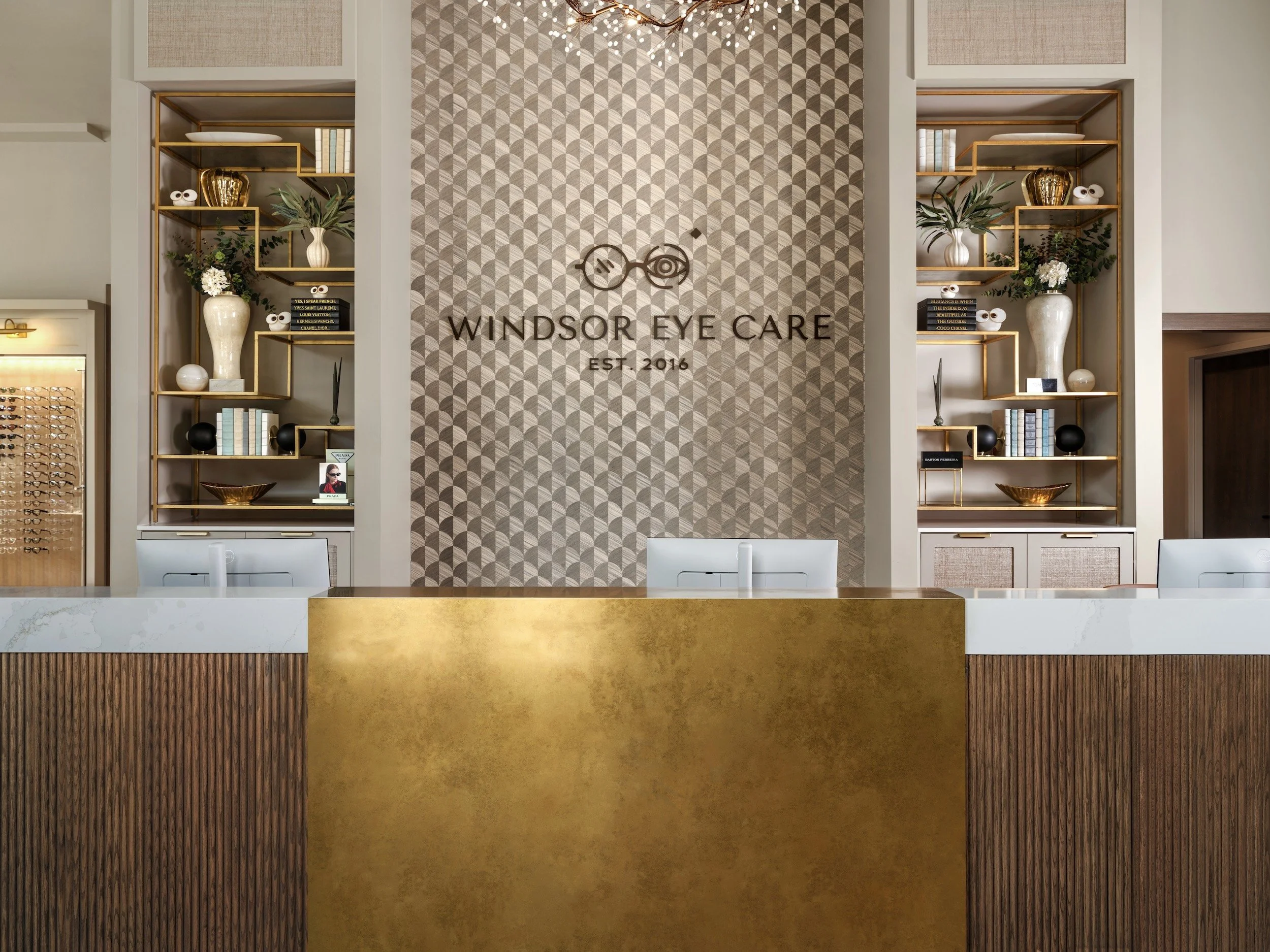Windsor Eye Care reception desk with gold-leaf panel, fluted walnut millwork, marble counter, and brass display shelving, designed by Luxe Style Studio
