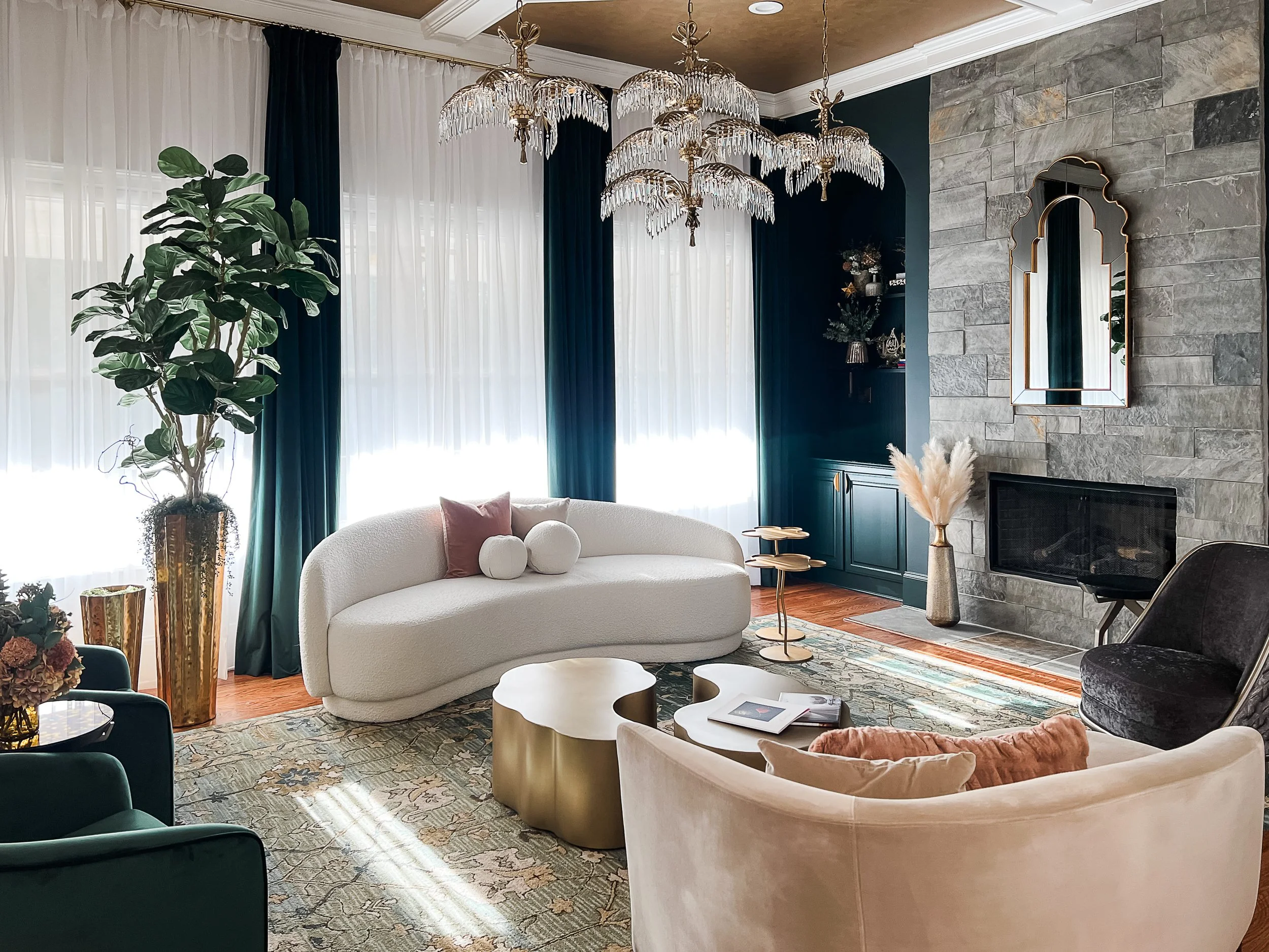 Living room with curved ivory seating, crystal chandeliers, dark green drapery, and a floor-to-ceiling stone fireplace, designed by Luxe Style Studio