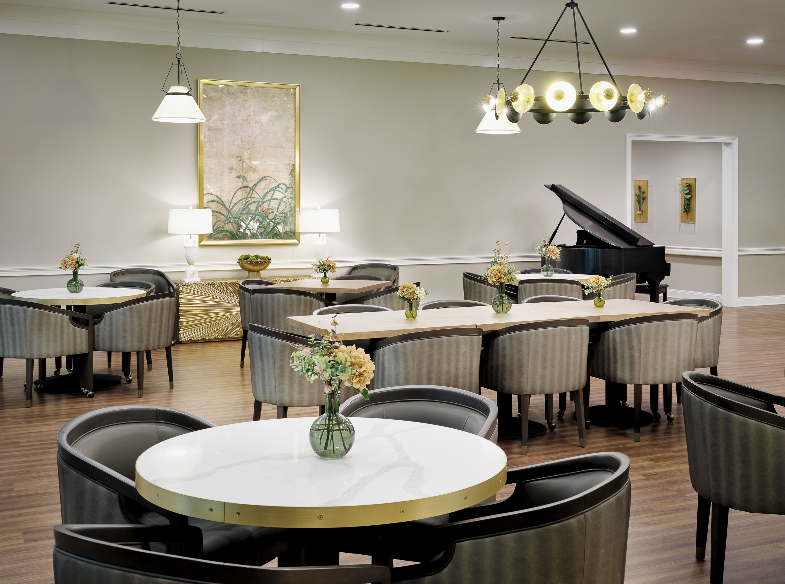 Dining room with grand piano, marble-top brass-edged round tables, barrel chairs, and sculptural gold chandelier, Thistle & Oak restaurant, Sugar Hill, GA, interior design by Ashley Schneider