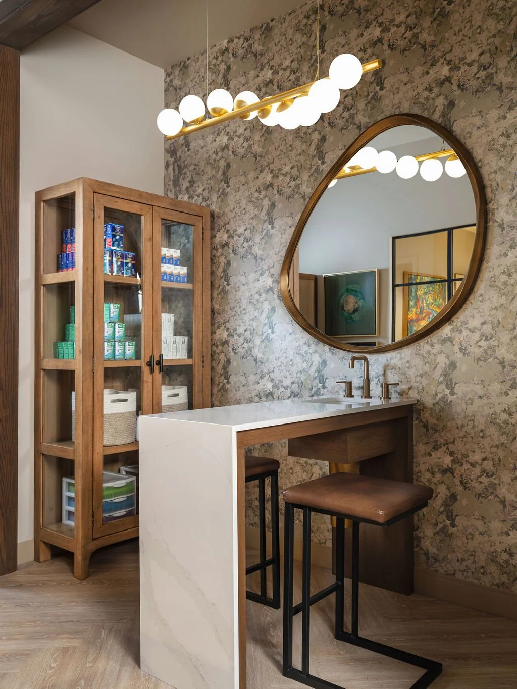 Boutique contact lens fitting room with botanical wallpaper, oversized round brass mirror, globe chandelier, white quartz counter, and wood display cabinet, interior design by Luxe Style Studio