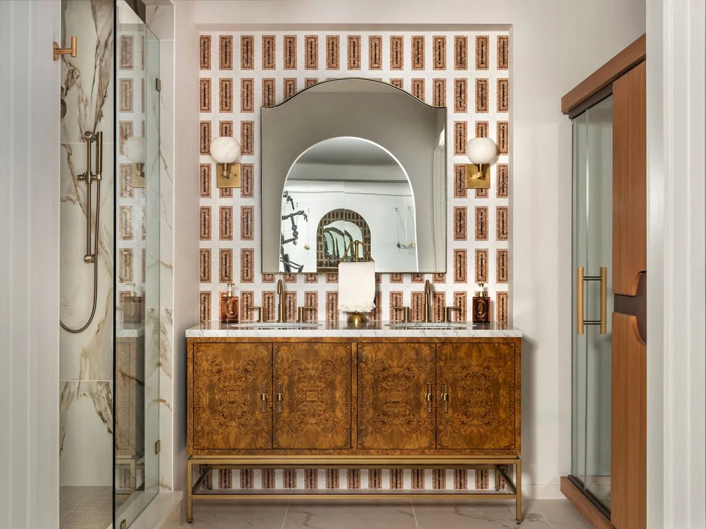 Home gym bathroom with burl wood vanity, marble countertop, arched mirror reflecting doorway beyond, geometric mosaic tile accent wall, infrared sauna, and brass globe sconces, interior design by Luxe Style Studio