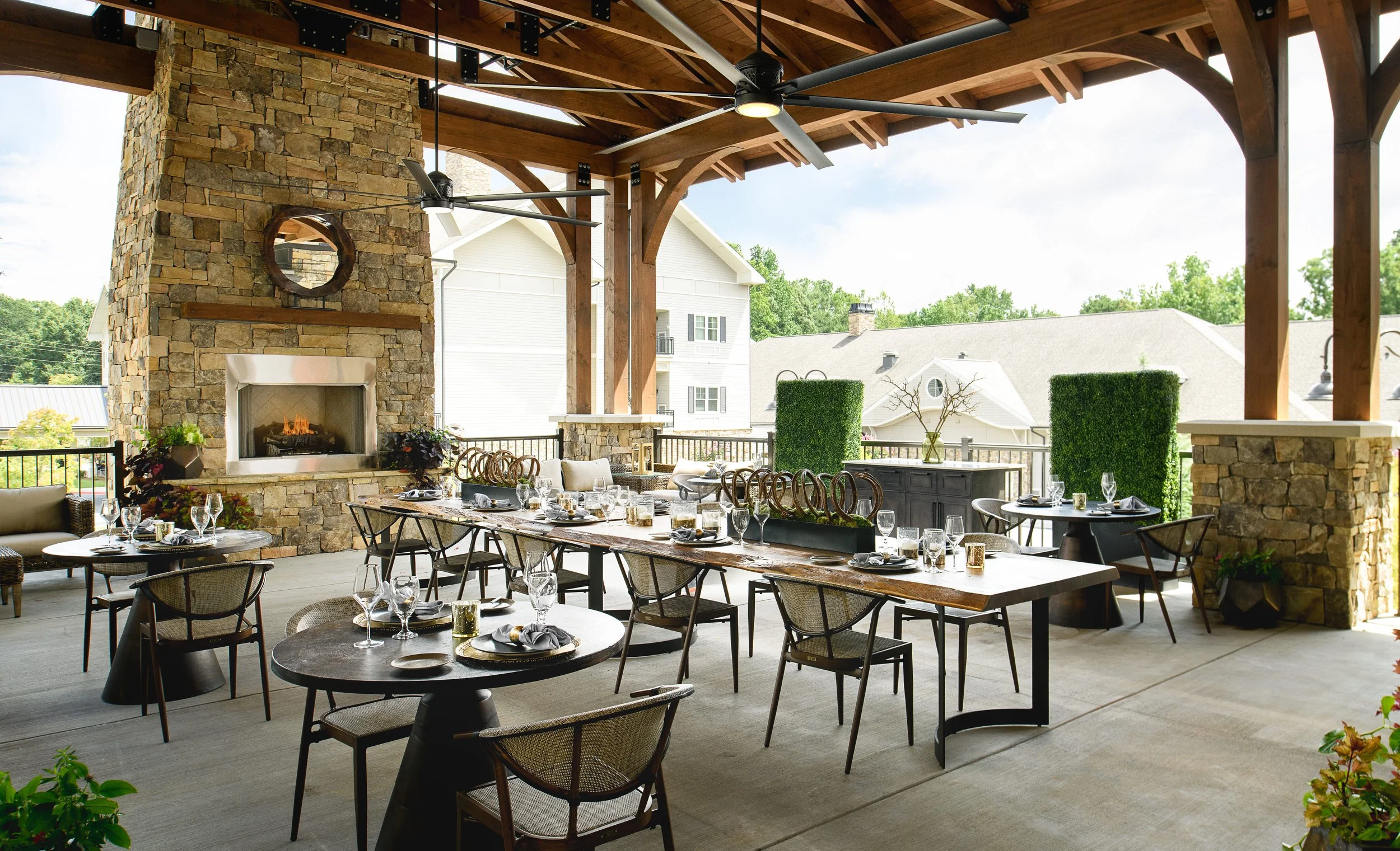 Local Hearth covered patio in Acworth with a timber pergola, stacked-stone fireplace, live-edge communal dining table, cane-back chairs, and boxwood panels, designed by Ashley Schneider