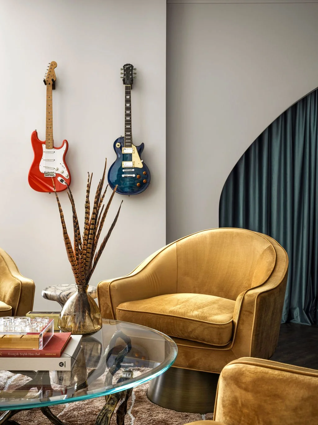 Eclectic music lounge with electric guitars mounted as wall art, gold velvet barrel chairs, sculptural glass coffee table, feather arrangement, and teal velvet arched drapes, interior design by Luxe Style Studio
