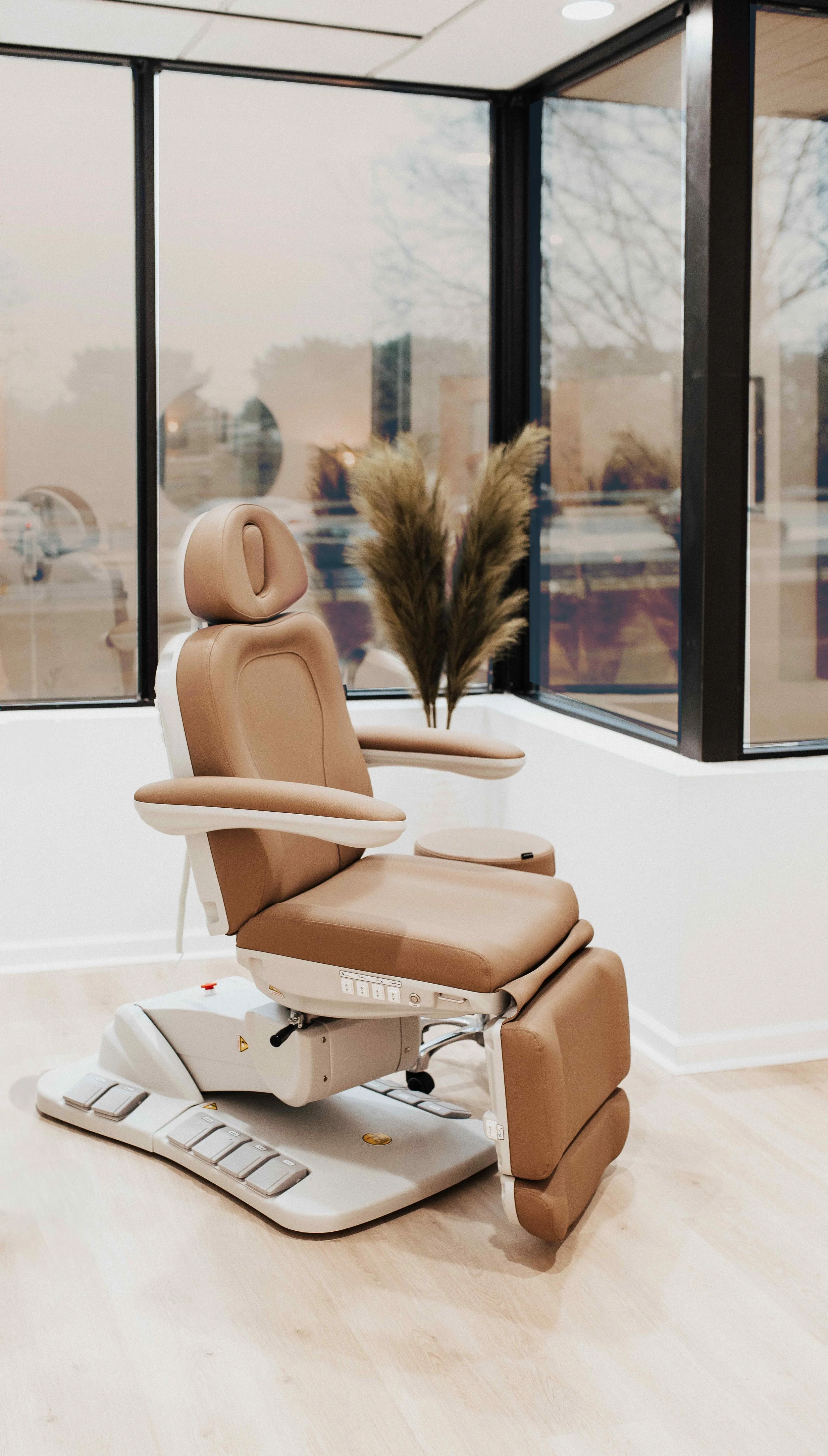 Luxury medical spa treatment chair in tan leather with white base positioned in bright corner room with floor-to-ceiling black steel windows and pampas grass arrangement, interior design by Luxe Style Studio