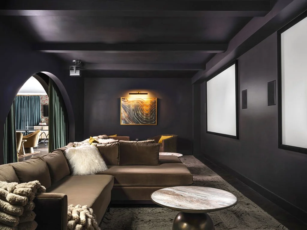 Luxury home theater with dark charcoal walls and ceiling beams, oversized velvet sectional, onyx side table, arched doorway, and dual large projection screens, interior design by Luxe Style Studio