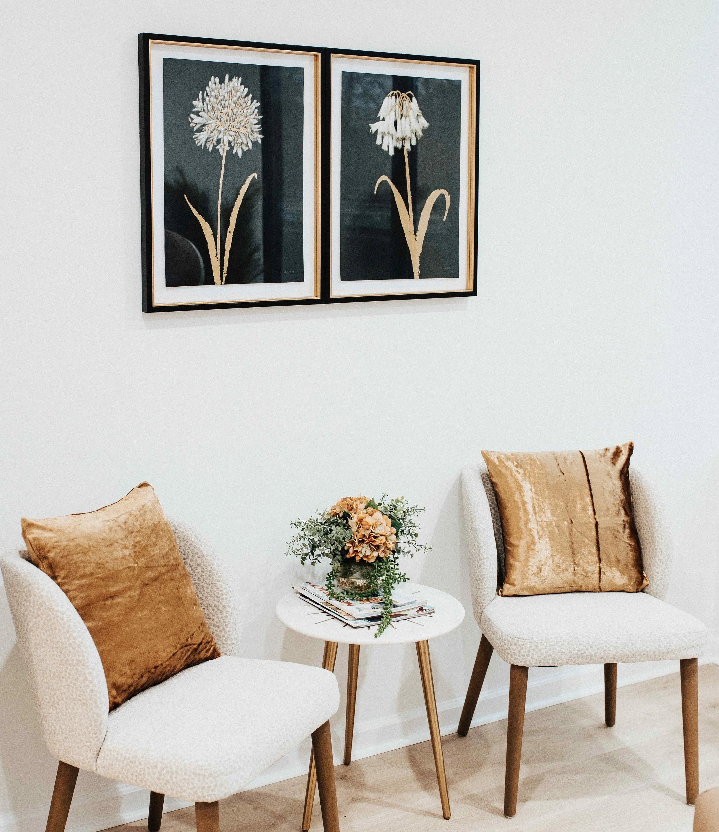 Intimate medical spa waiting area with two cream bouclé accent chairs, gold velvet pillows, white marble side table, and paired botanical artwork on dark background with gold frames, interior design by Luxe Style Studio