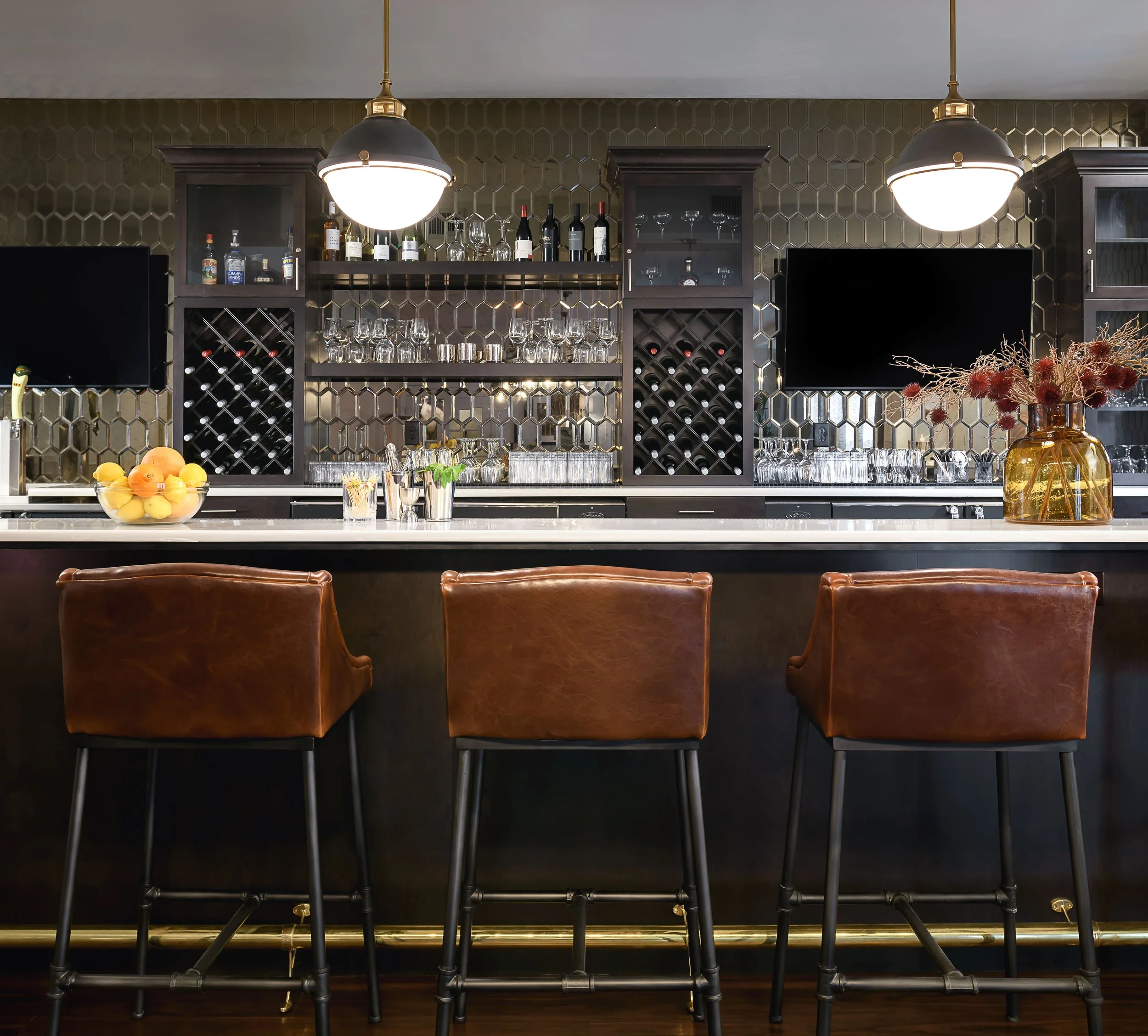 Local Hearth bar in Acworth with floor-to-ceiling black honeycomb tile, dark wood cabinetry, wine storage, a pale stone counter, cognac leather stools, and brass pendant lights, designed by Ashley Schneider