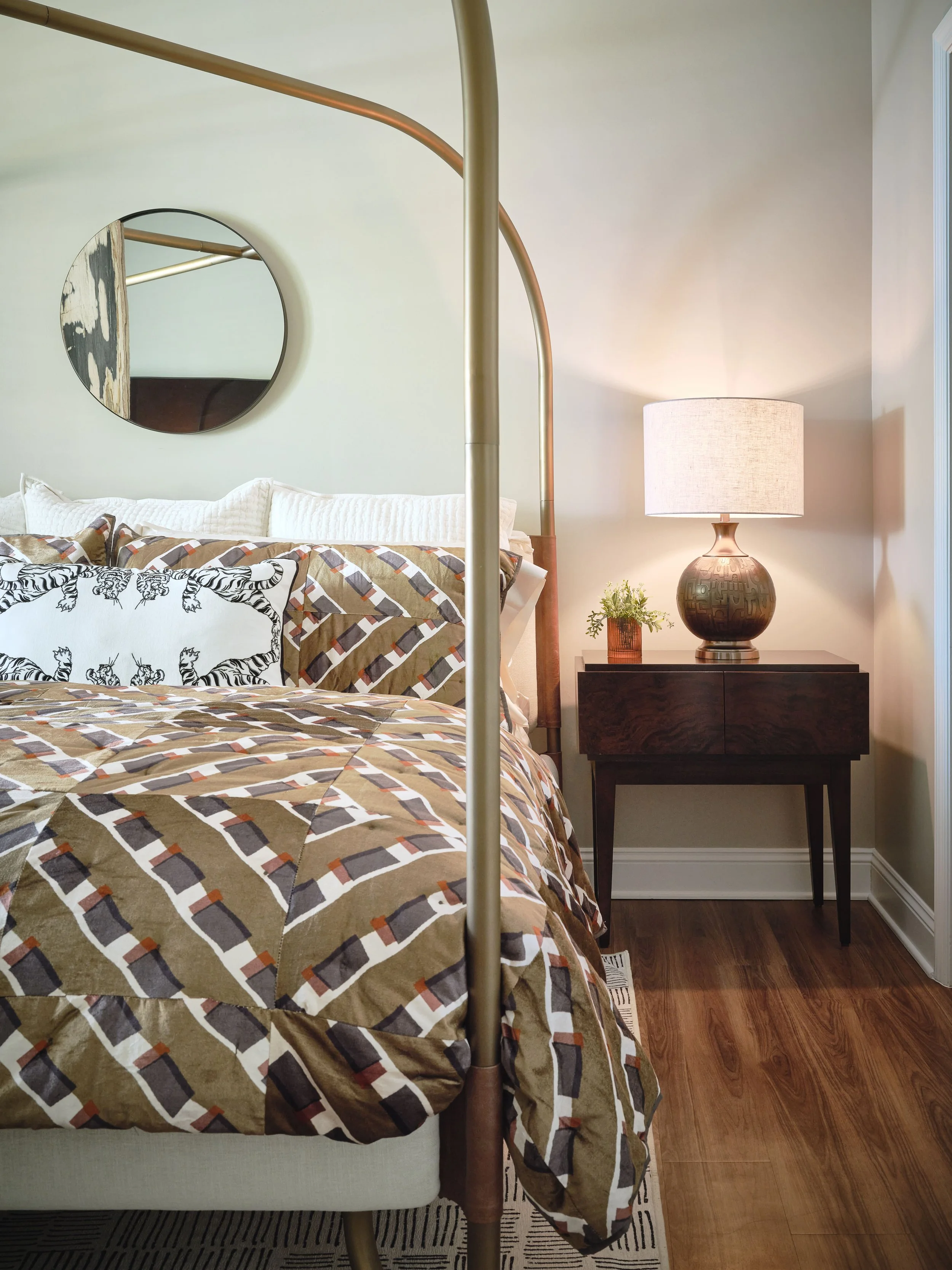 West Broad bedroom detail with a dark walnut mid-century nightstand, textured brass ceramic lamp, round abstract mirror, brass canopy-bed post, and geometric bedding, designed by Ashley Schneider