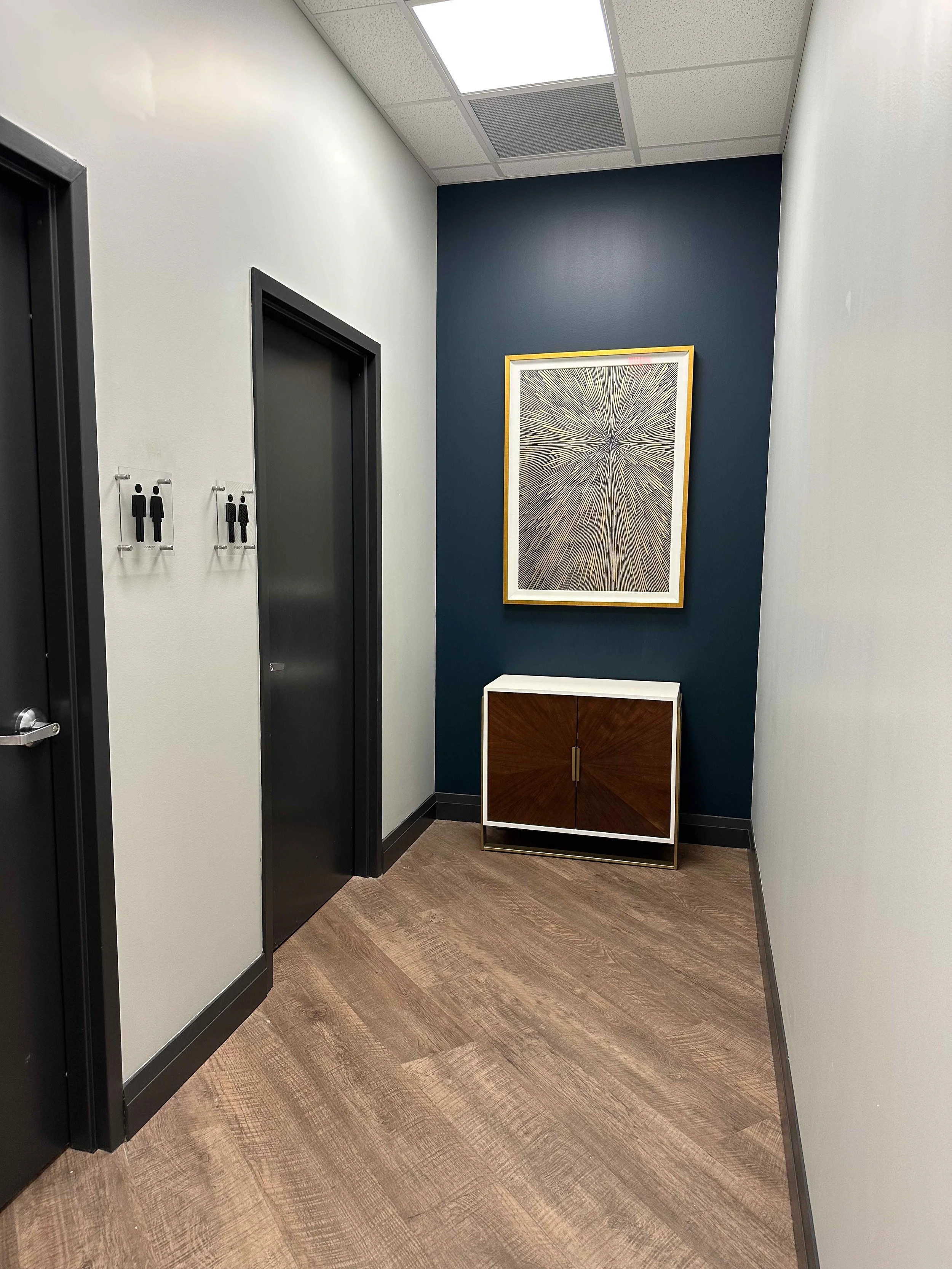 Corporate office restroom corridor with navy blue accent wall, large starburst artwork in gold frame, sunburst wood accent cabinet with white marble top, and dark framed doors, interior design by Luxe Style Studio