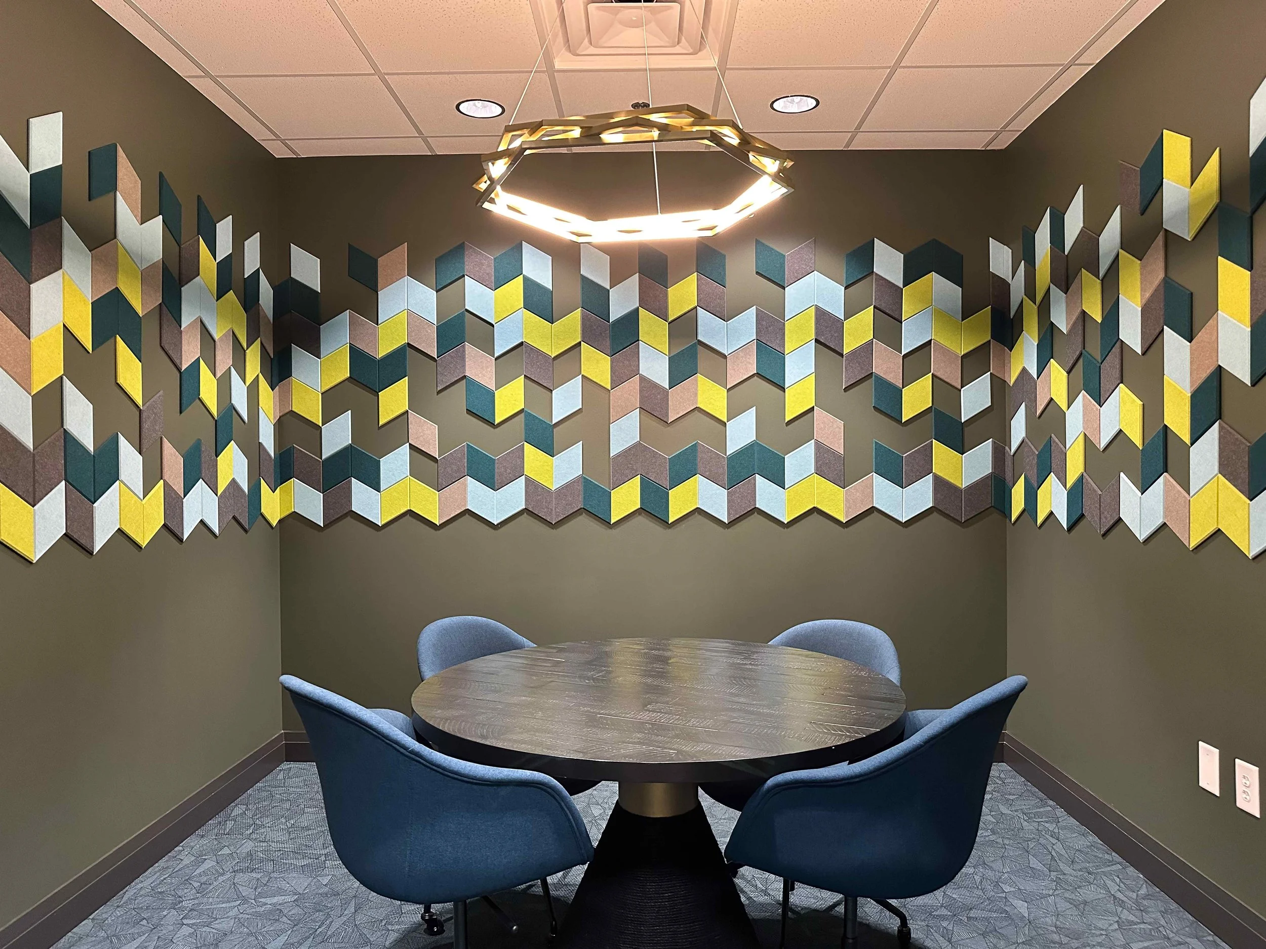 Corporate conference room with floor-to-ceiling colorful geometric acoustic wall panels in teal, yellow, and mauve, round dark wood table, blue upholstered chairs, and hexagonal brass chandelier, interior design by Luxe Style Studio