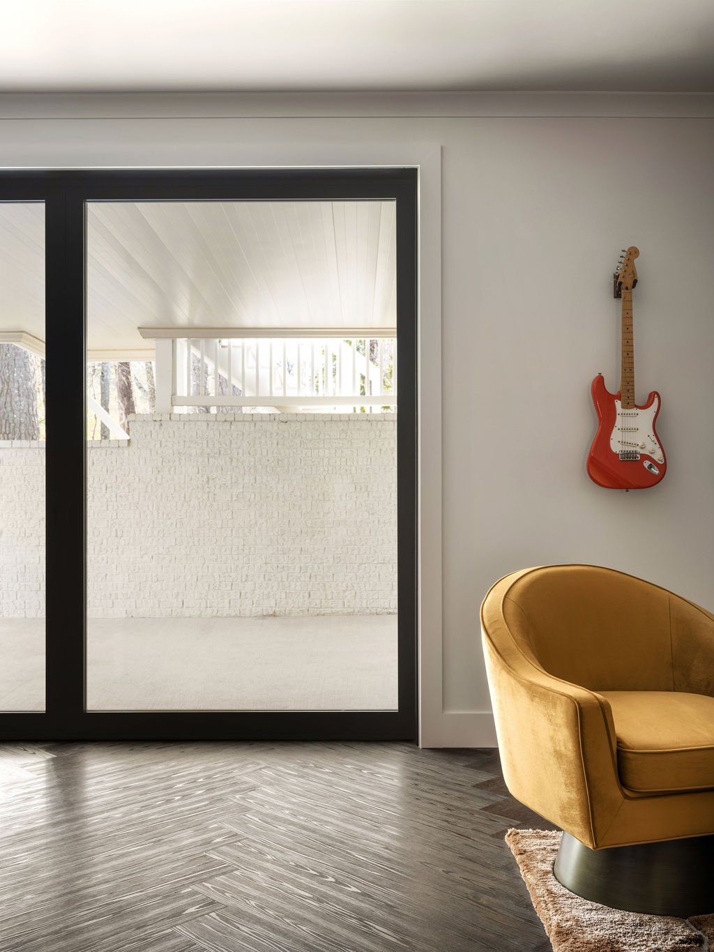 Eclectic lounge with red electric guitar mounted as wall art, gold velvet swivel chair, dark herringbone floors, and oversized black steel sliding glass door to covered porch, interior design by Luxe Style Studio