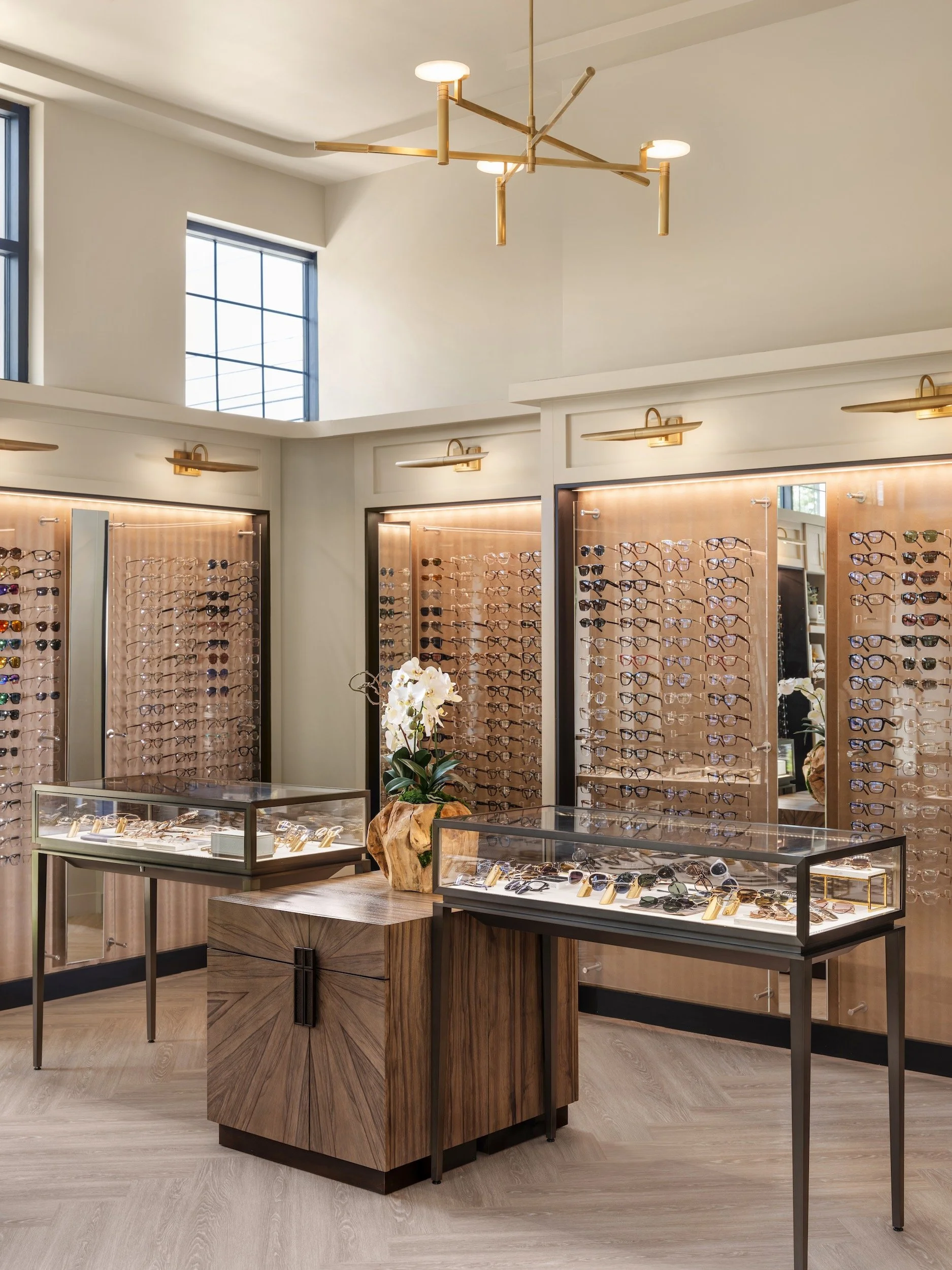 Luxury optical boutique interior with illuminated wood eyewear display walls, glass showcase tables, sunburst wood island, and brass sputnik chandelier, Atlanta, interior design by Luxe Style Studio