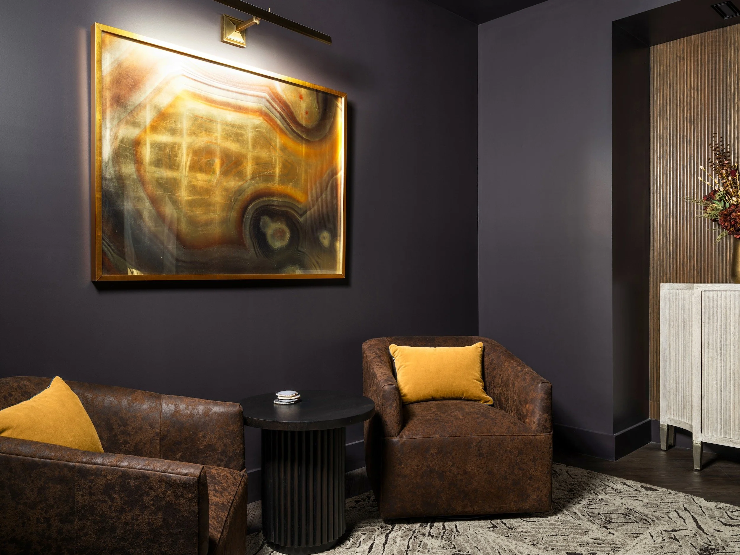 Moody sitting room with leather chairs, amber artwork, and deep aubergine walls, designed by Luxe Style Studio