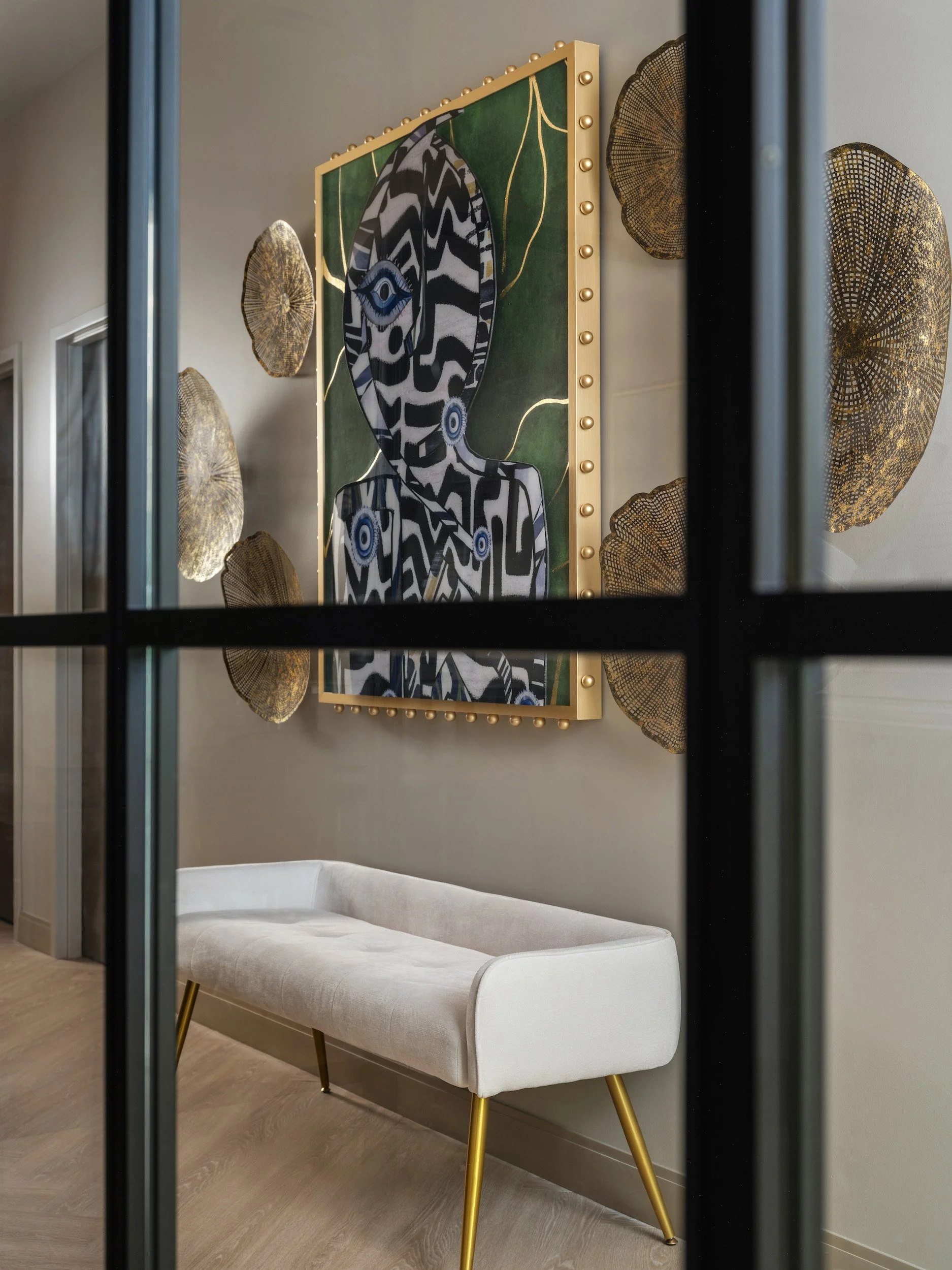 Art vignette through a black steel partition with bold figurative painting, gold fan wall sculptures, and cream velvet bench, Windsor Eye Care, Atlanta, GA, interior design by Luxe Style Studio