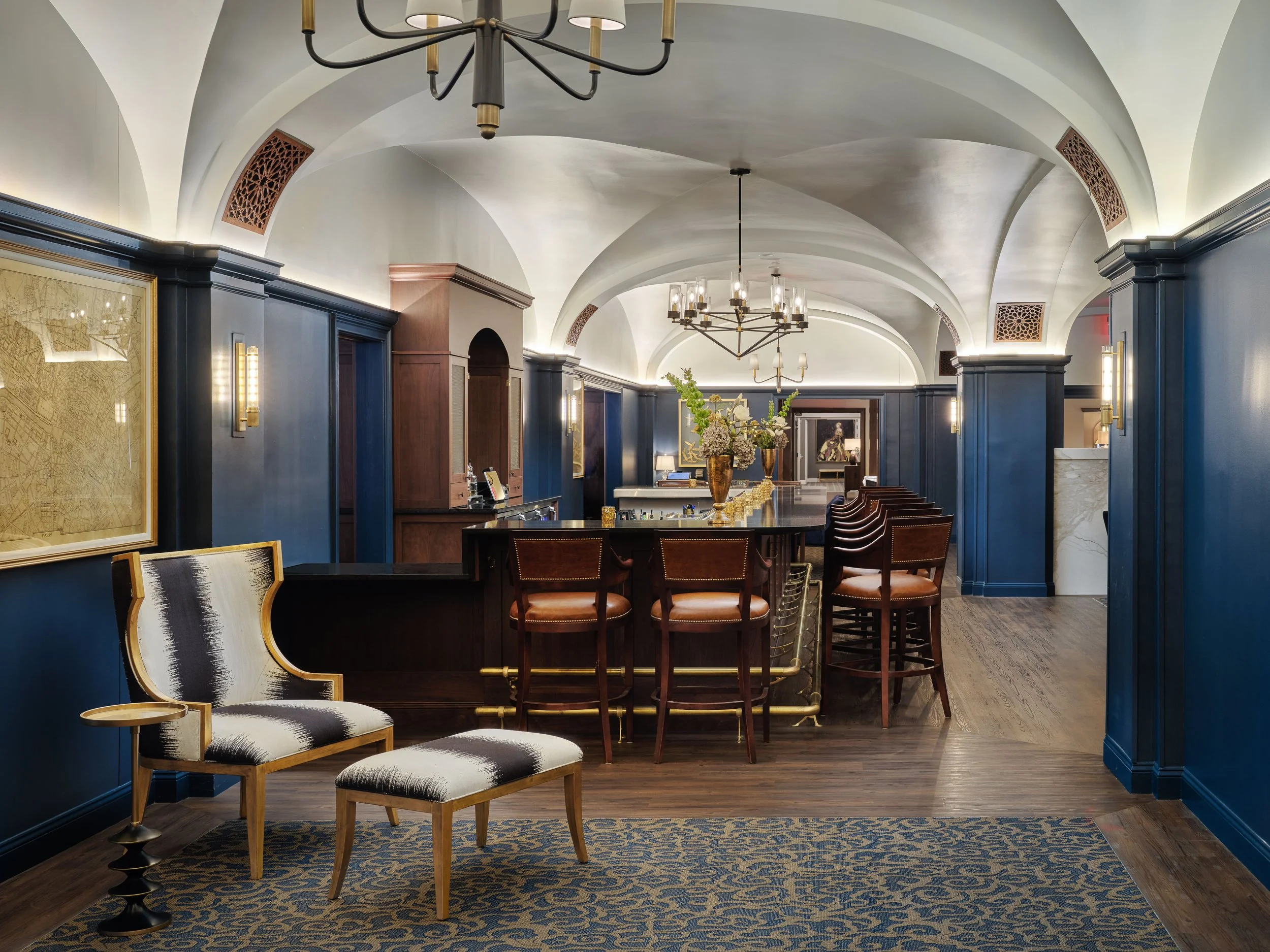 Off The Rails bar with a vaulted ceiling, navy millwork, cognac leather stools, brass lighting, and a black-and-white accent chair, designed by Ashley Schneider