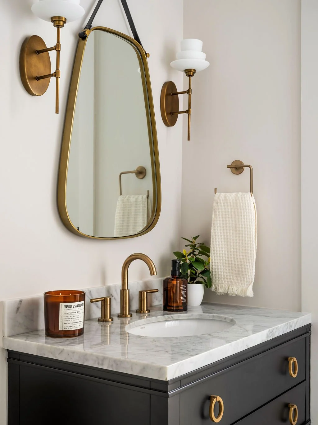 Powder room with matte black vanity, white marble countertop, brass ring hardware, leather strap brass-framed mirror, and globe brass sconces, interior design by Luxe Style Studio