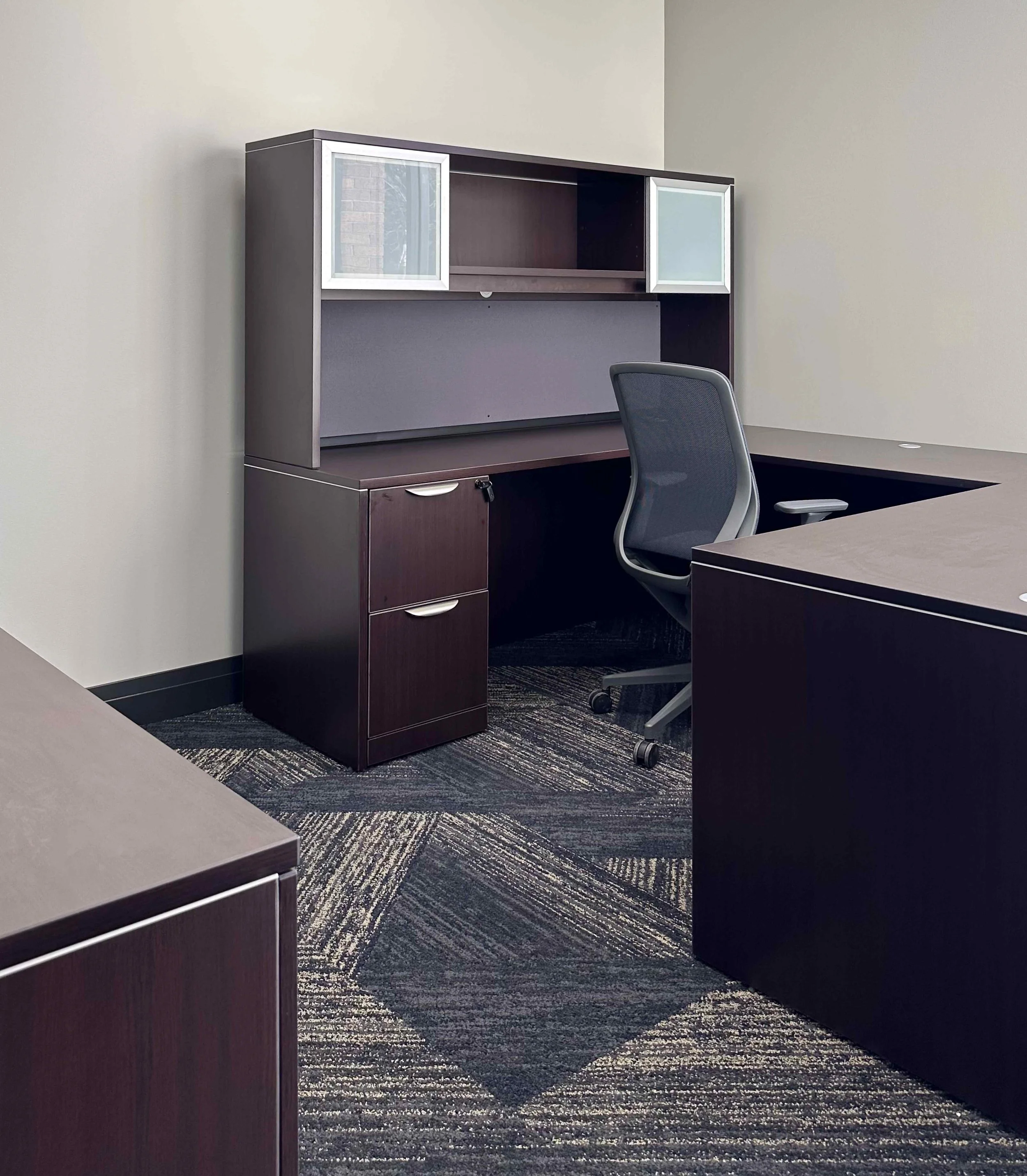 Private corporate office workstation with dark espresso L-shaped desk, overhead storage hutch with frosted glass doors, mesh task chair, and geometric patterned carpet tile, interior design by Luxe Style Studio