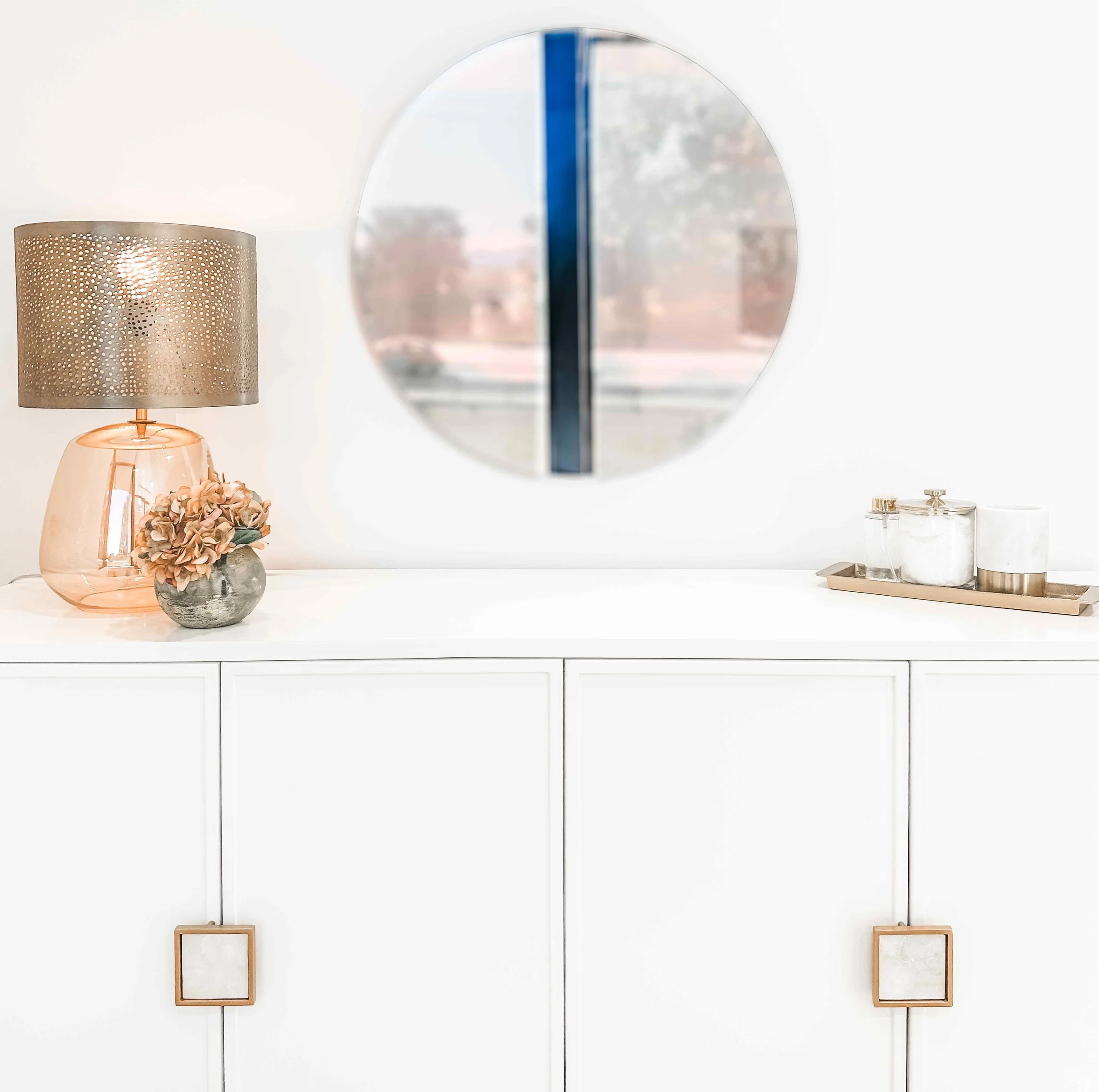 Luxury spa treatment room detail with white shaker cabinet, square brass hardware, amber glass table lamp with perforated shade, round wall mirror, and delicate floral arrangement, interior design by Luxe Style Studio
