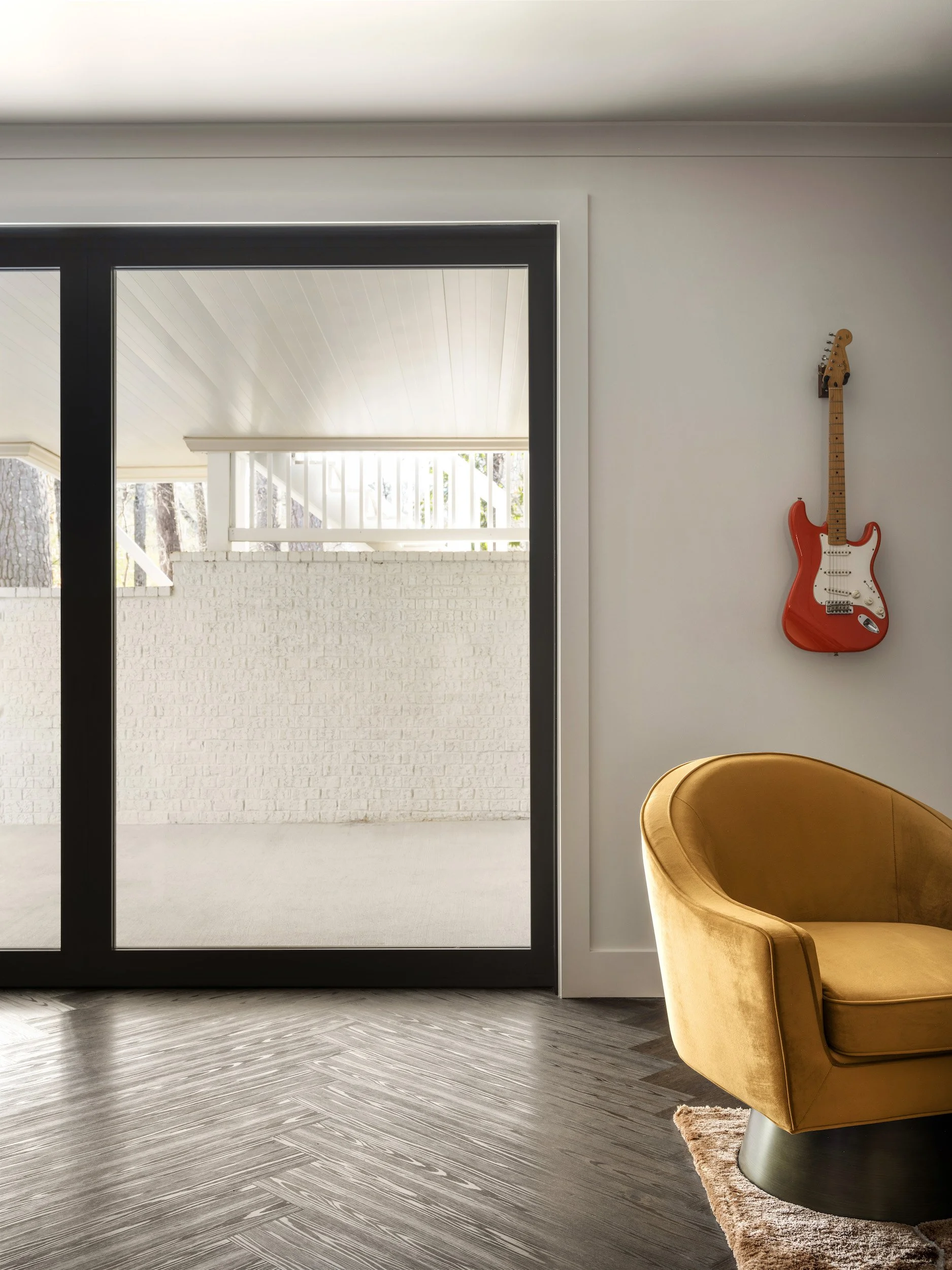 Eclectic lounge with red electric guitar mounted as wall art, gold velvet swivel chair, dark herringbone floors, and oversized black steel sliding glass door to covered porch, Atlanta, GA private residence, interior design by Luxe Style Studio