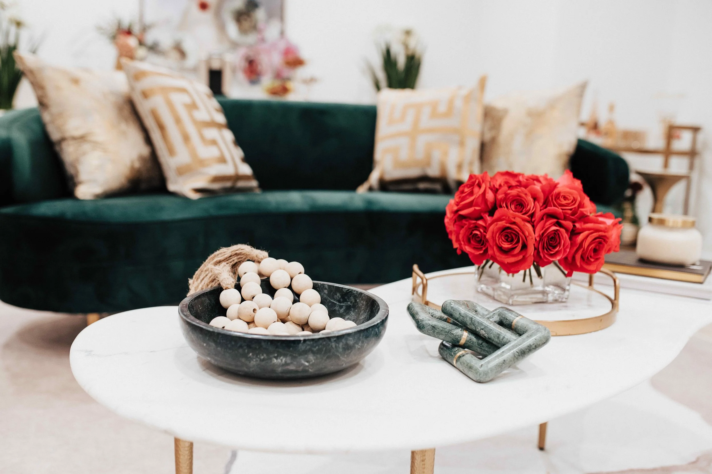 Luxury medical spa coffee table detail with marble top, dark stone bowl, red rose arrangement on brass tray, and green marble sculpture, with curved emerald velvet sofa and gold Greek key pillows beyond, interior design by Luxe Style Studio