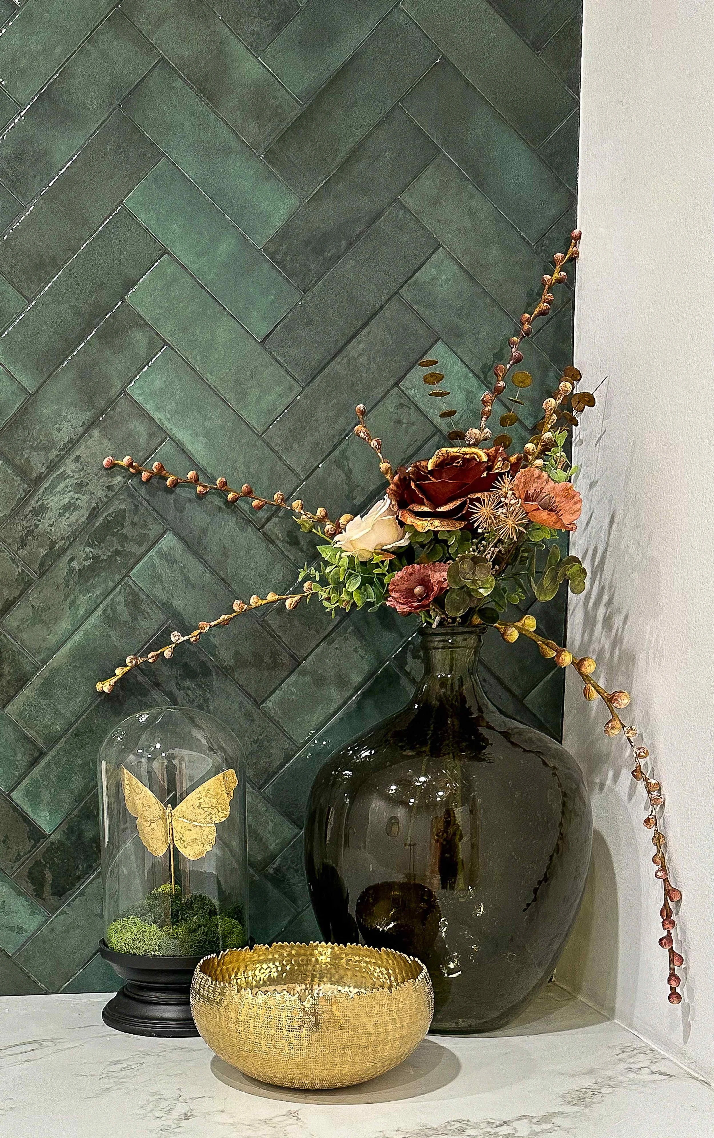Luxury spa accent wall detail with deep green herringbone zellige tile, large dark glass vase with botanical arrangement, gold butterfly cloche, and hammered brass bowl, interior design by Luxe Style Studio