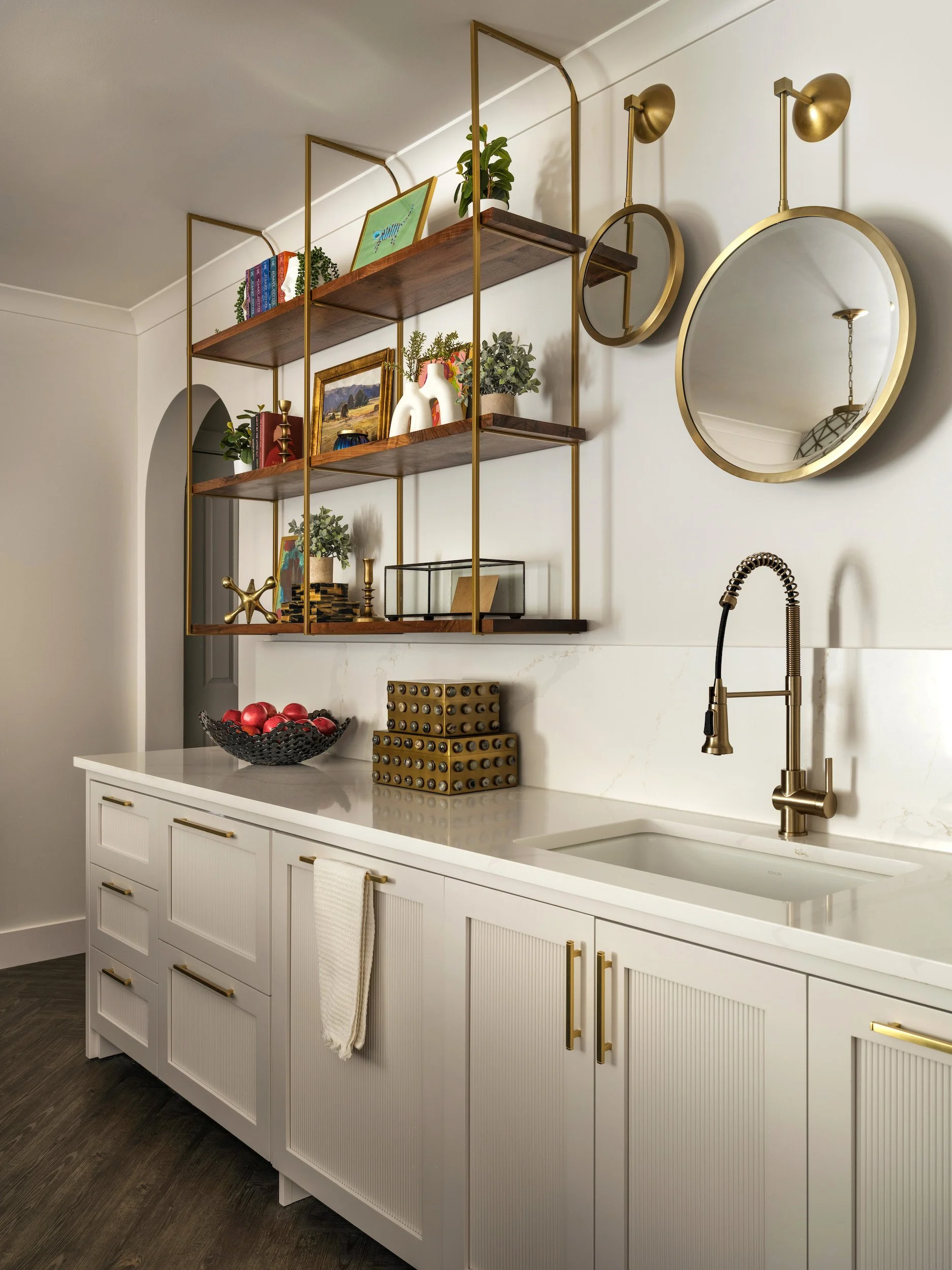 White fluted wet bar cabinetry with brass shelving, marble backsplash, and a brass faucet, designed by Luxe Style Studio