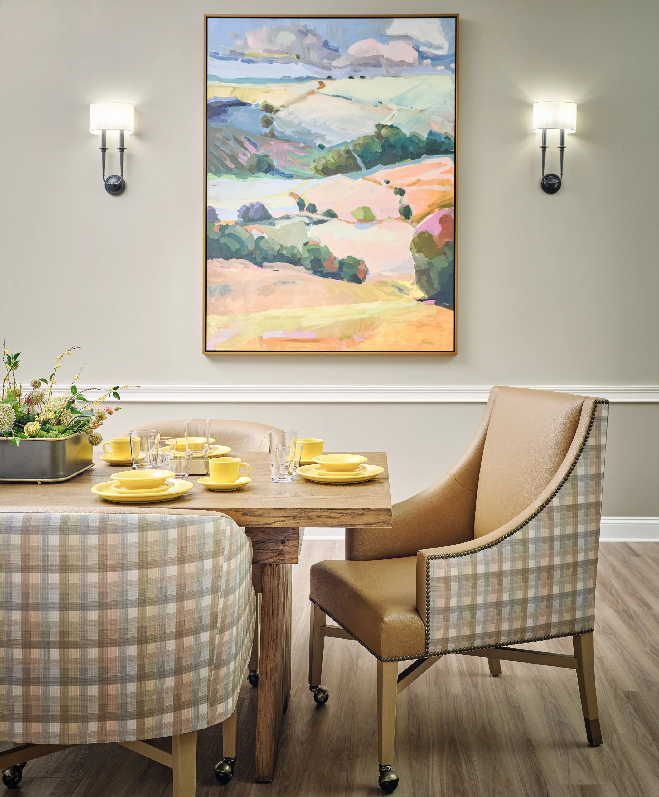 Thistle & Oak dining nook with plaid chairs, a camel leather host chair, yellow tableware, landscape artwork, and black sconces, designed by Ashley Schneider