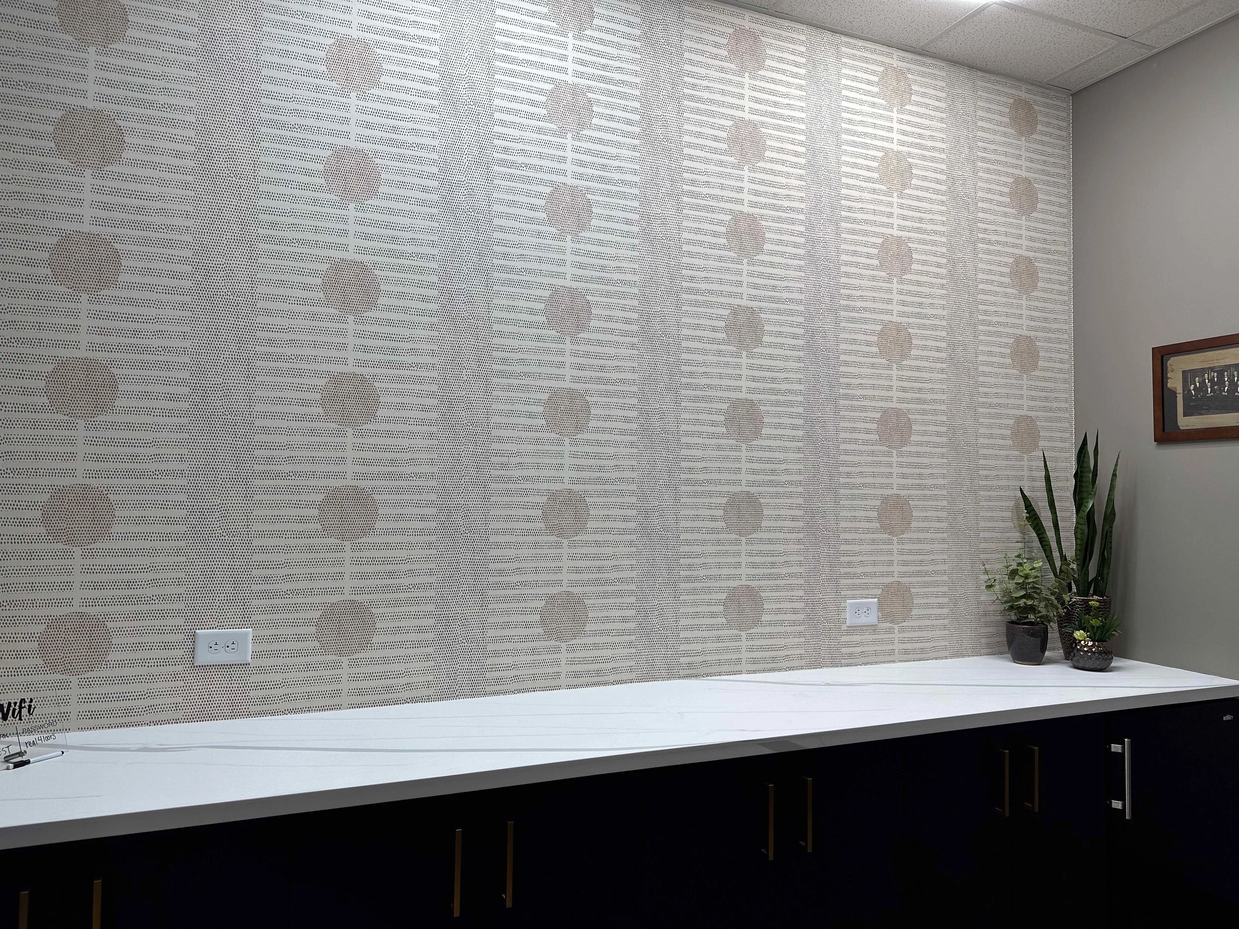 Corporate training room with large-scale neutral geometric textured wallpaper accent wall, white quartz countertop, dark base cabinets with brass hardware, and snake plant styling, interior design by Luxe Style Studio