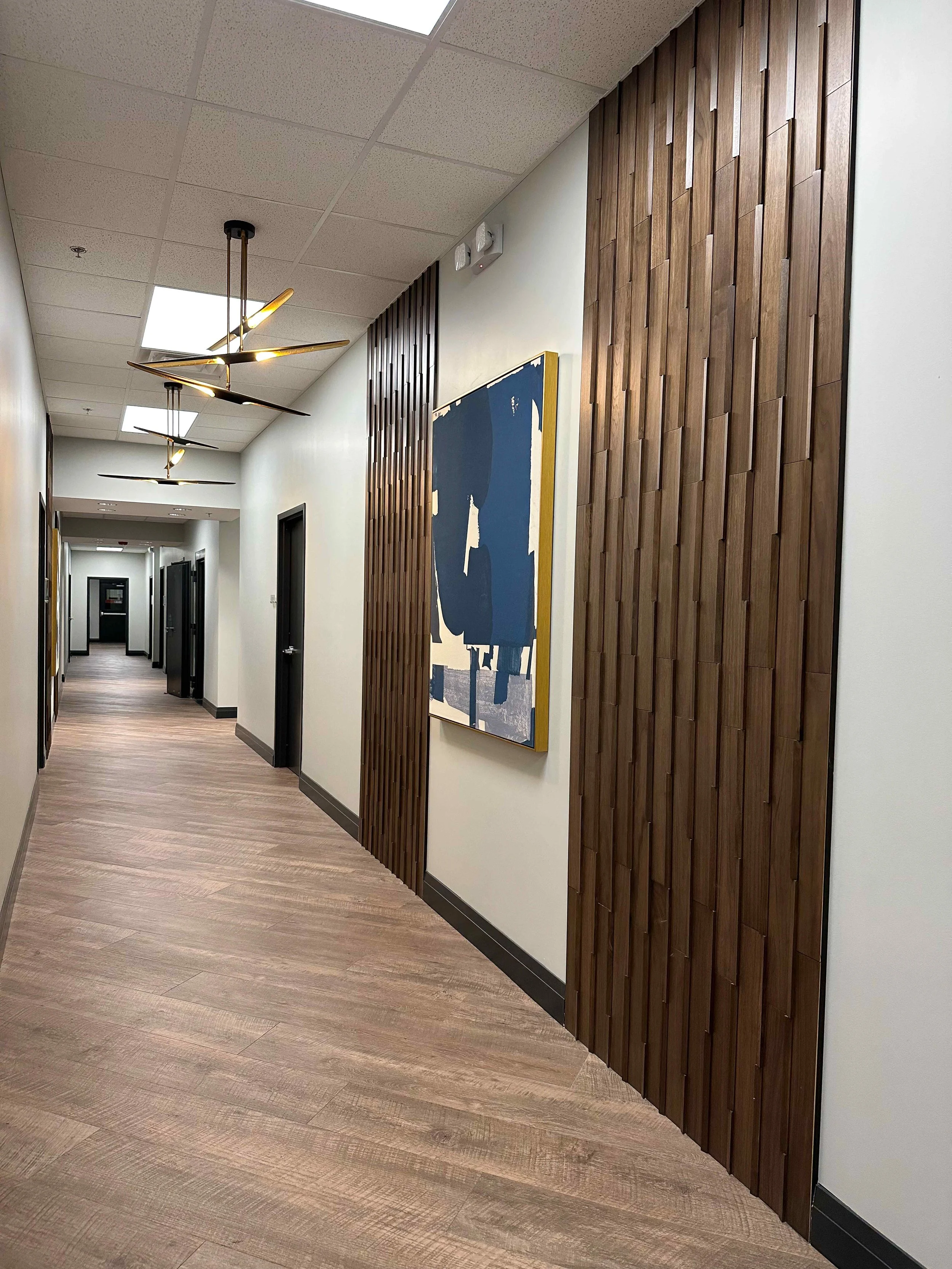 Corporate office hallway with floor-to-ceiling walnut wood slat accent wall, large abstract blue and gold framed artwork, brass angular pendant lights, and wood plank floors leading to private offices, interior design by Luxe Style Studio