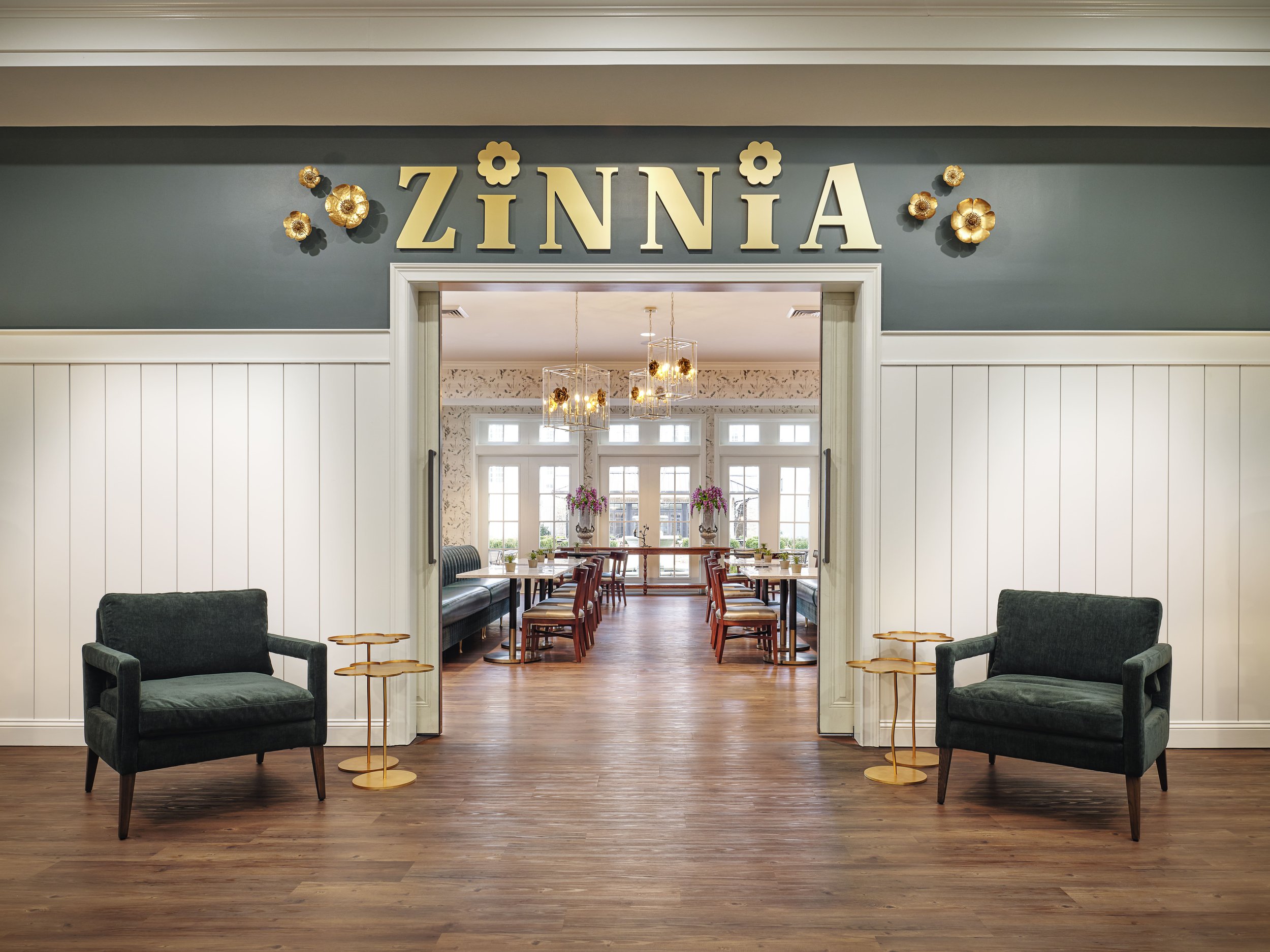 Zinnia Café & Market entrance with gold dimensional signage, floral wall sculptures, green velvet armchairs, and a view into the dining room, designed by Ashley Schneider