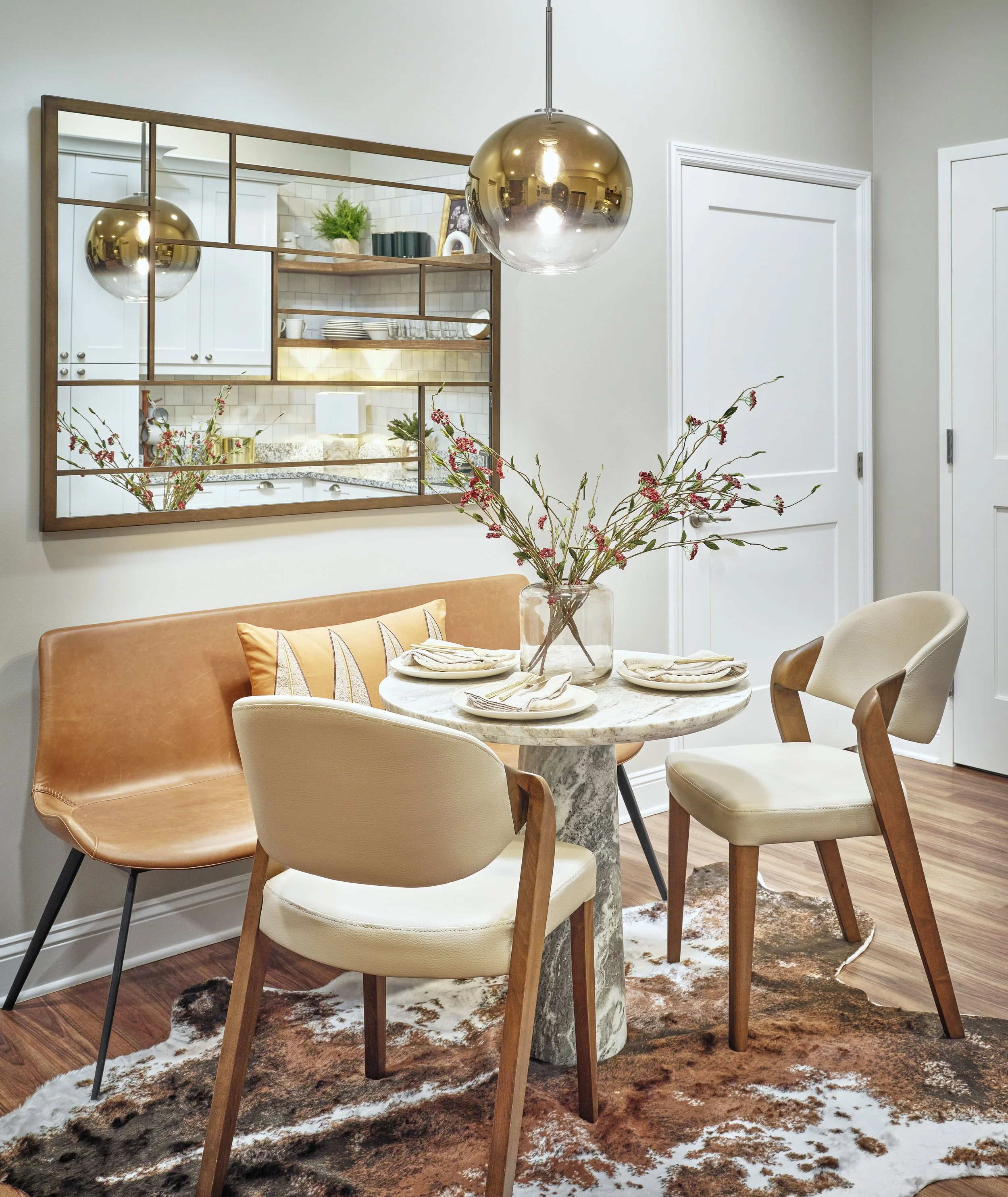 West Broad dining nook in Sugar Hill with a round marble pedestal table, tan leather bench, cream upholstered chairs, brass ombre globe pendant, geometric brass mirror, cowhide rug, and cherry blossom centerpiece, designed by Ashley Schneider