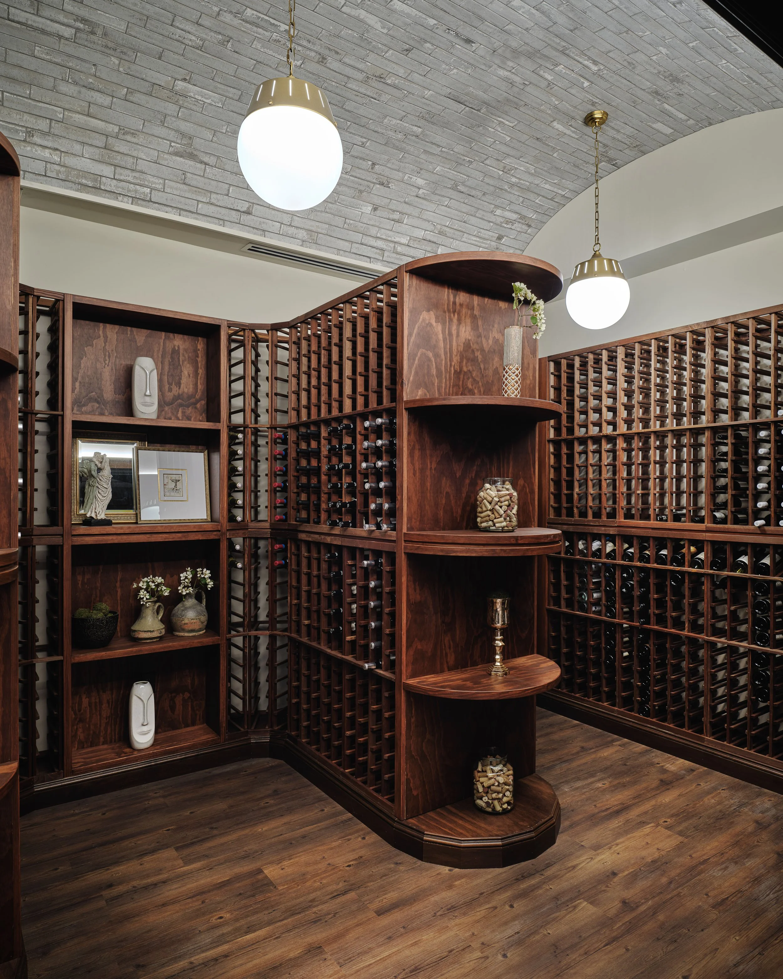 Custom walnut wine cellar with floor-to-ceiling bottle storage, curved display shelving, a brick-vaulted ceiling, and globe pendants at Off The Rails, designed by Ashley Schneider
