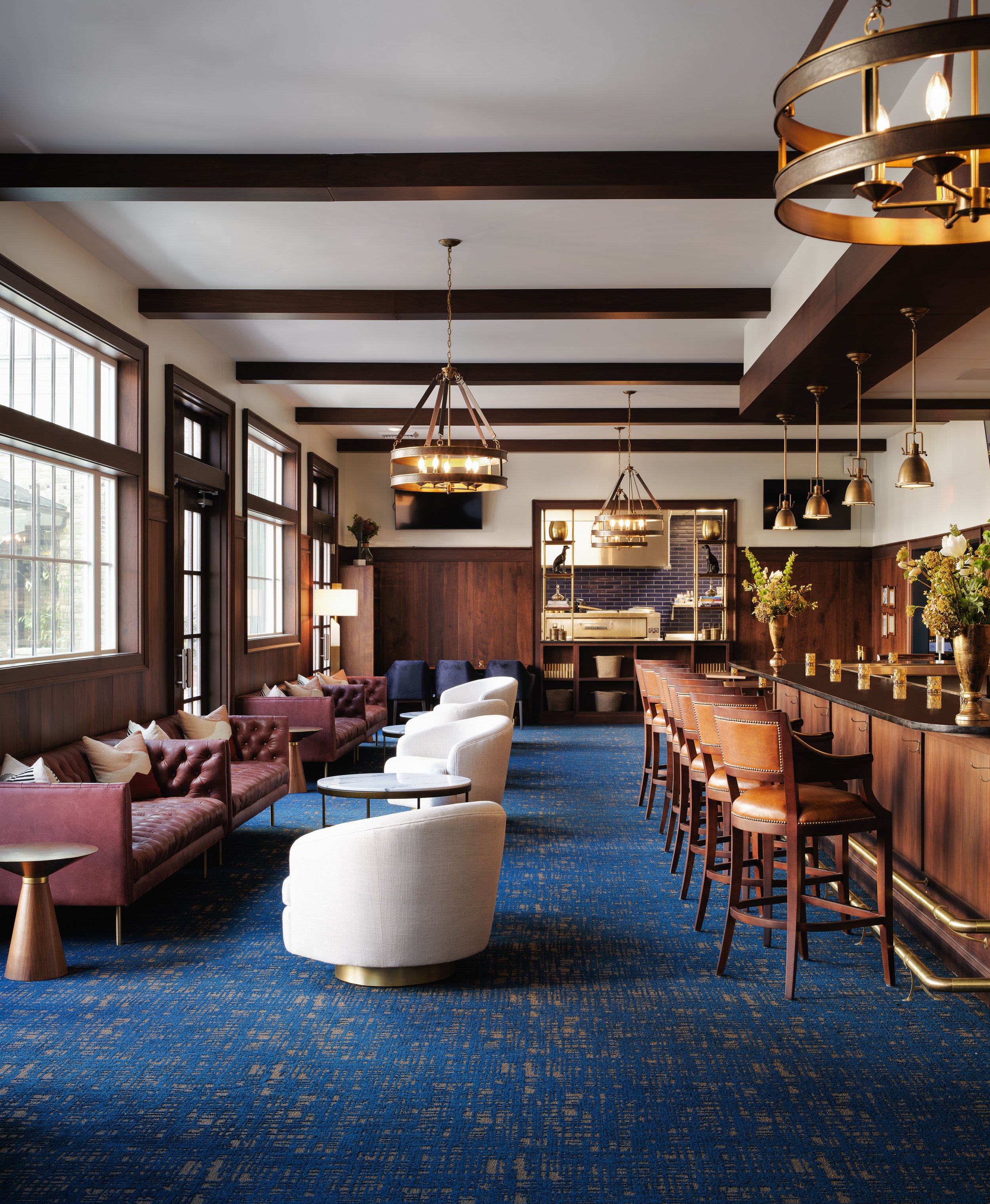 Off The Rails lounge with a burgundy tufted sofa, white swivel chairs, cognac leather bar stools, wood beams, and brass chandeliers, designed by Ashley Schneider