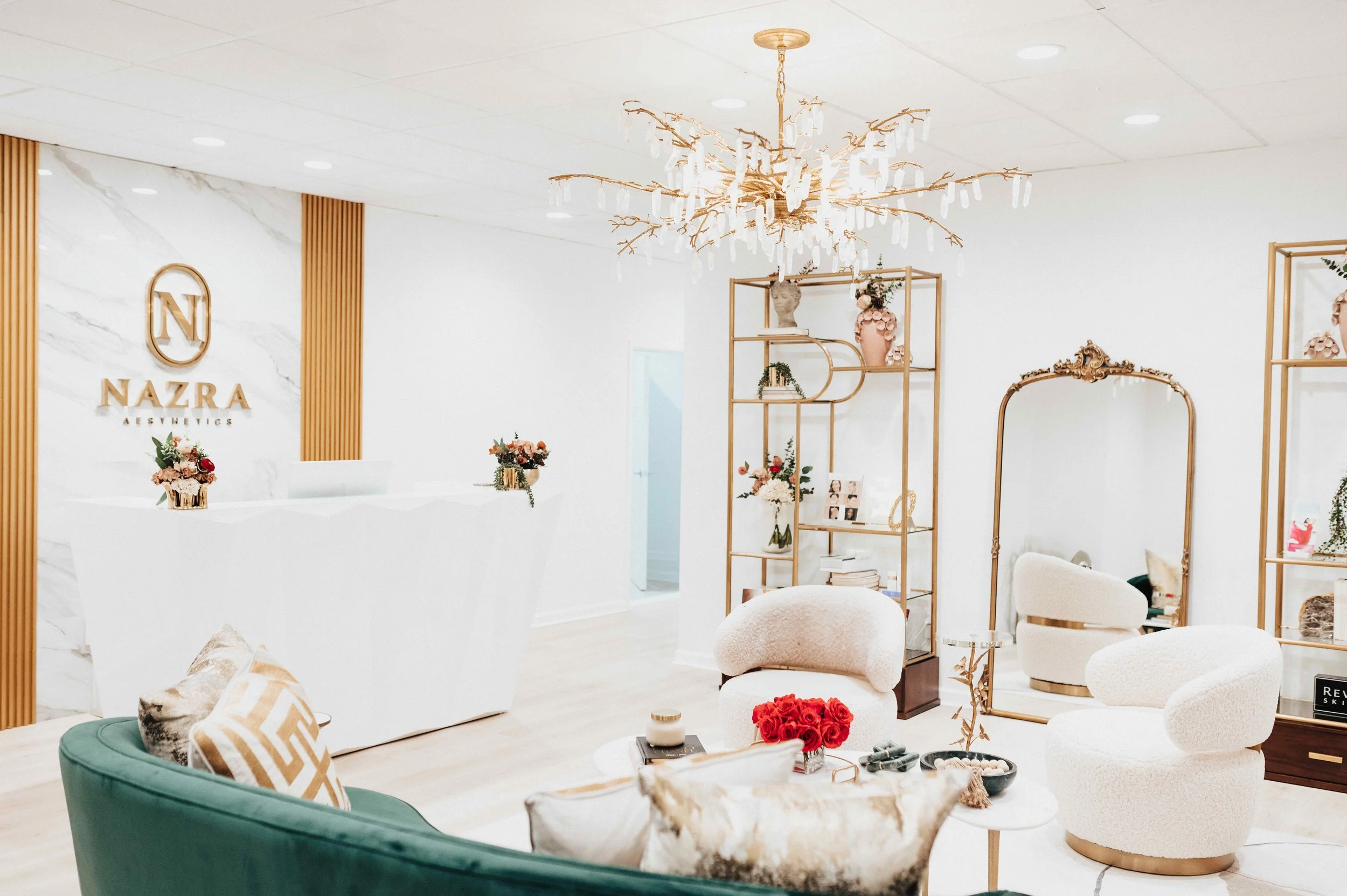 Full view of Nazra Aesthetics reception area with white marble reception desk, gold logo signage, fluted wood accent wall, branch crystal chandelier, gold art deco etageres, and ornate floor mirror, interior design by Luxe Style Studio