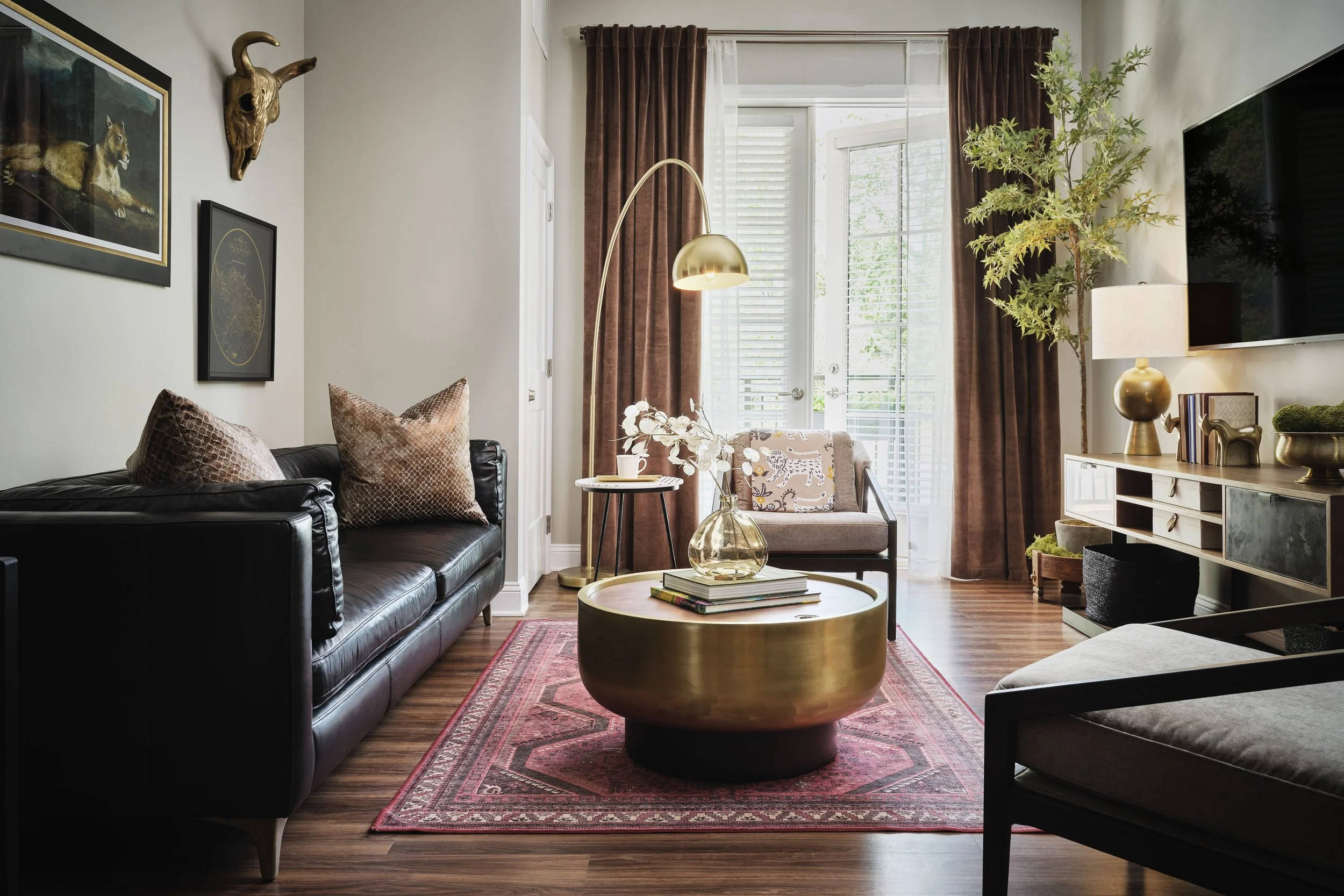 Eclectic living room wide view with black leather sofa, oversized brass drum coffee table, brass arc floor lamp, brown velvet drapes, vintage Persian rug, gold bull skull wall mount, and animal art gallery wall, interior design by Ashley Schneider