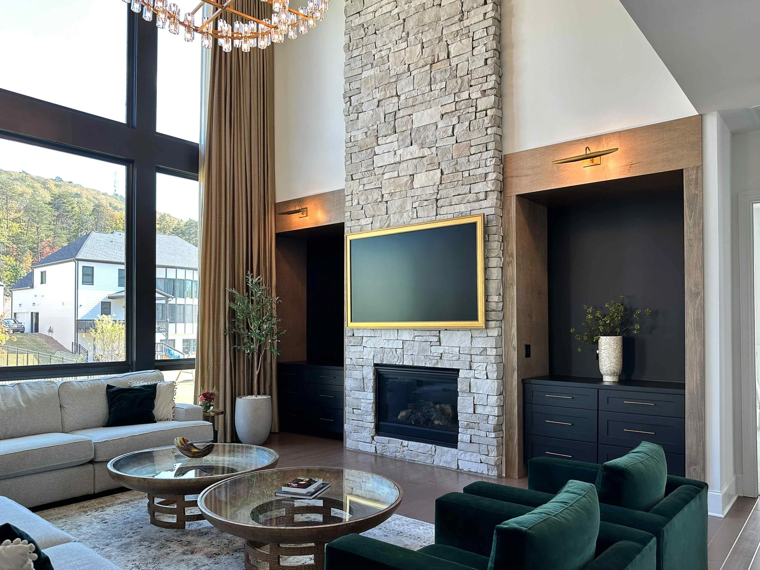 Grand great room featuring floor-to-ceiling stacked stone fireplace, dark built-in cabinetry, and emerald velvet accent chairs by Luxe Style Studio
