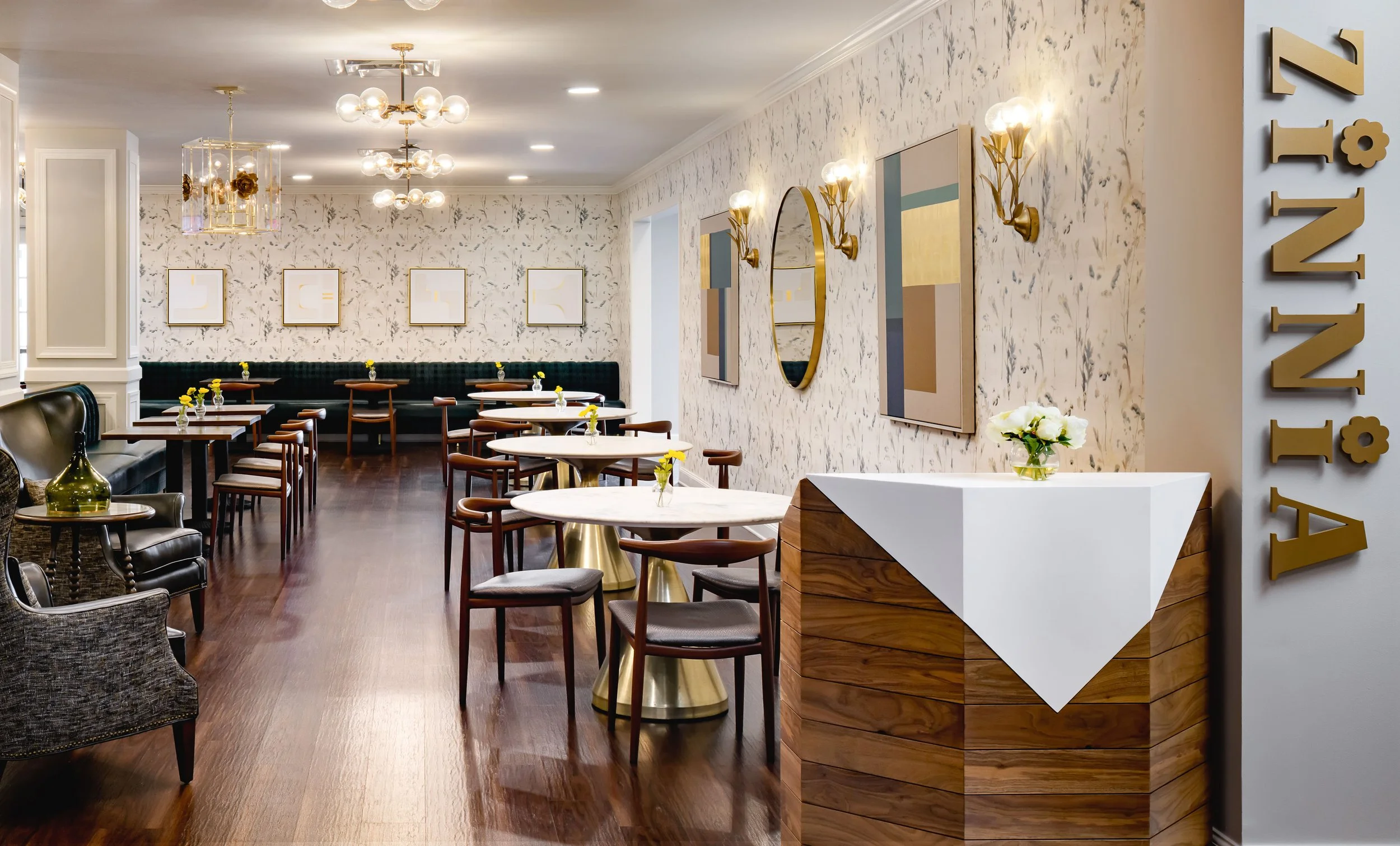 Zinnia Parisian-inspired café host stand and full dining room with marble pedestal tables, walnut chairs, green leather banquette, botanical wallpaper, brass globe chandelier, and sculptural walnut host stand, interior design by Ashley Schneider