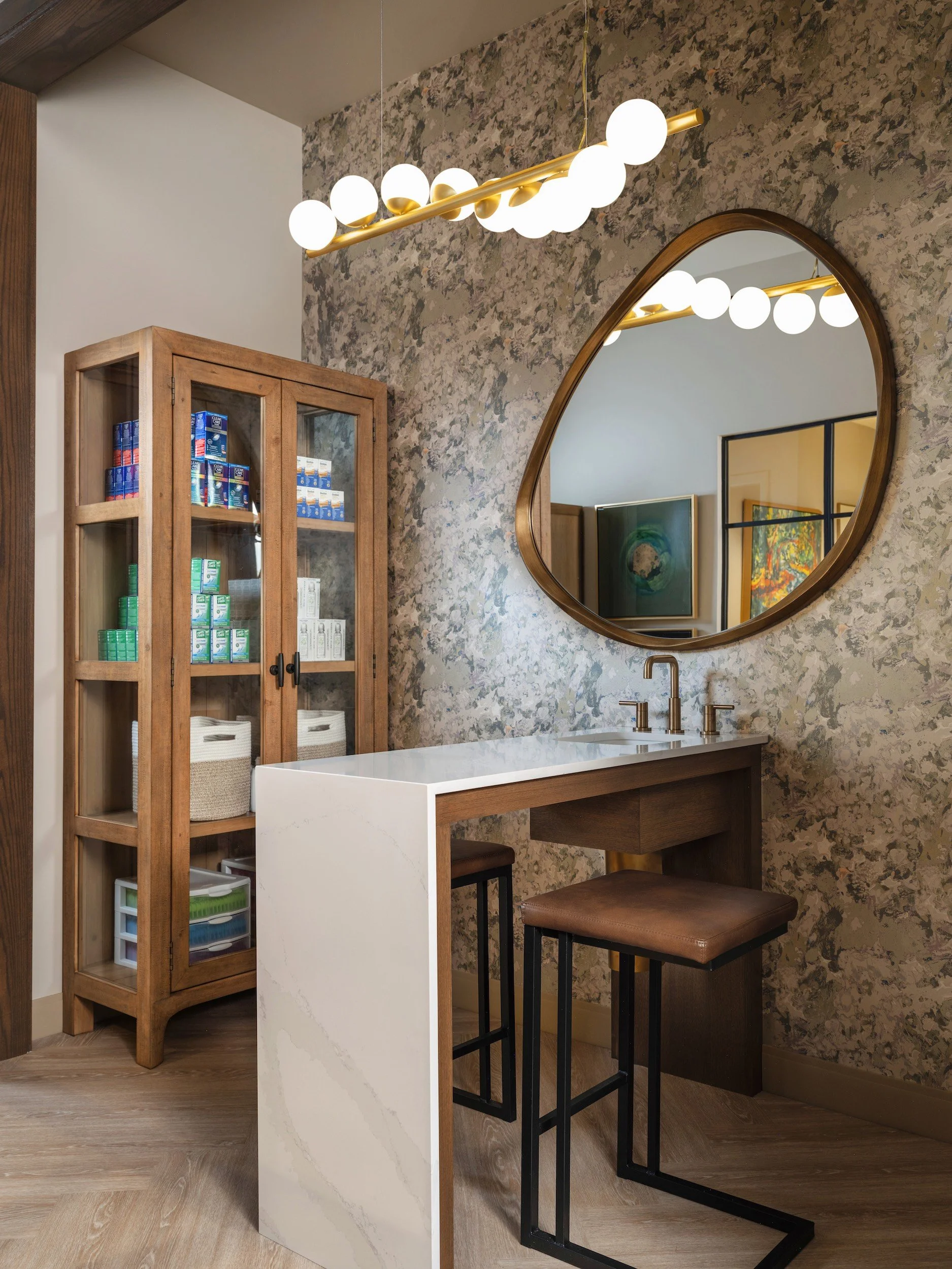 Boutique contact lens fitting room with botanical wallpaper, oversized round brass mirror, globe chandelier, white quartz counter, and wood display cabinet, Windsor Eye Care optical boutique, Atlanta, GA, interior design by Luxe Style Studio