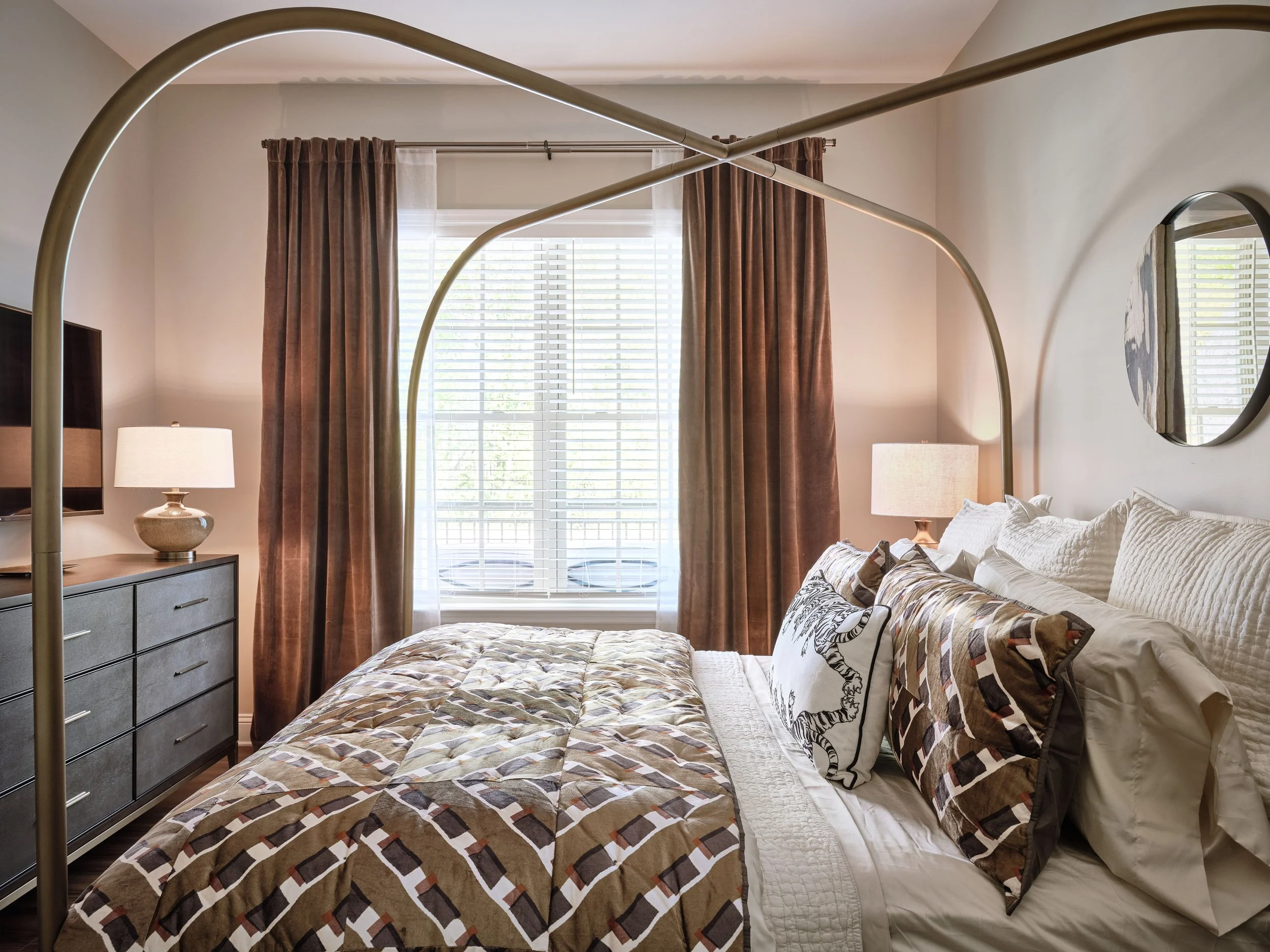 West Broad primary bedroom in Sugar Hill with a sculptural brass canopy bed, geometric bedding in olive and brown, brown velvet drapery, ceramic table lamps, a round wall mirror, and a mud-cloth accent rug, designed by Ashley Schneider