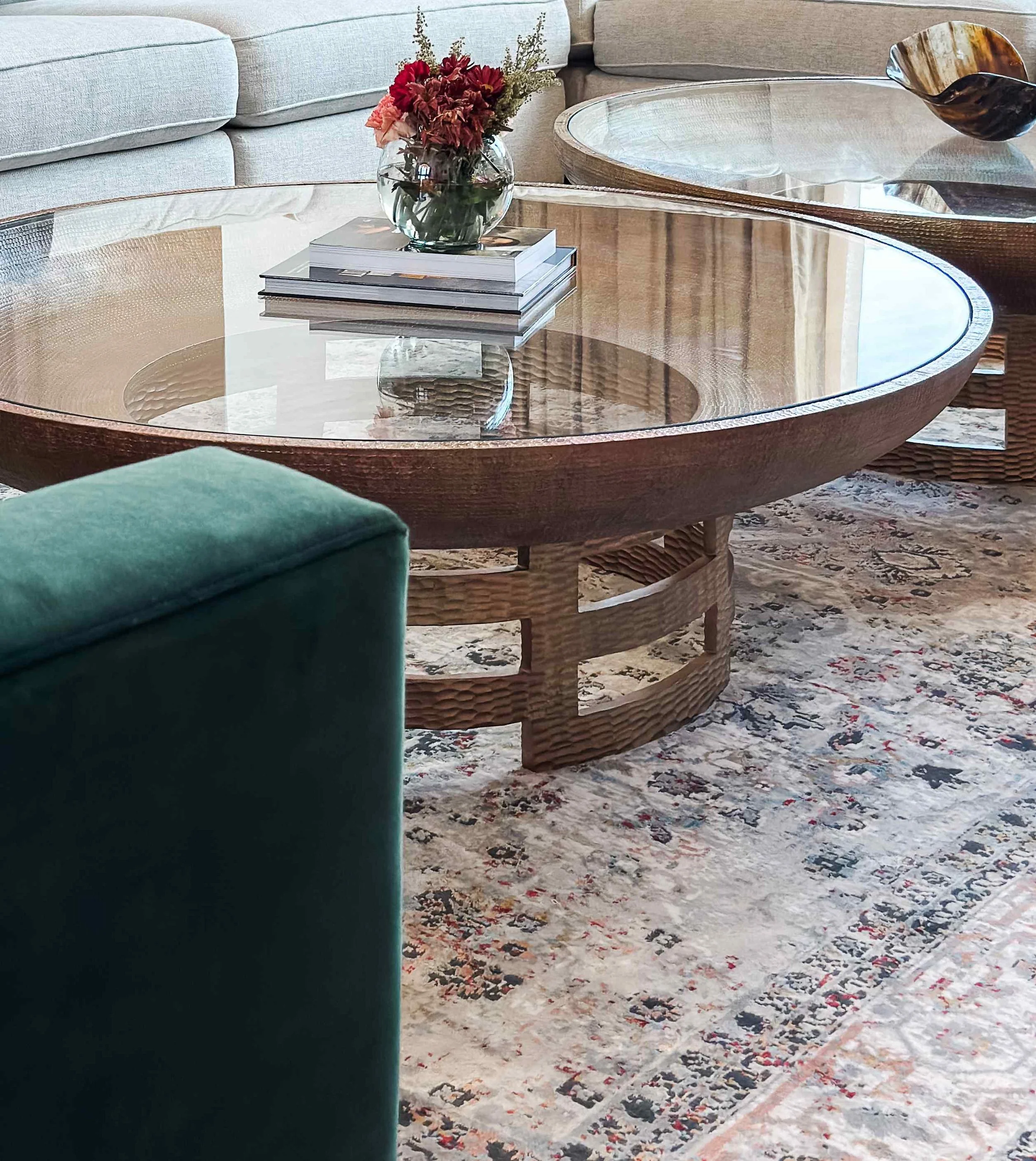 Close-up of hammered bronze and glass round coffee table on Persian-style area rug with emerald velvet ottoman, Luxe Style Studio
