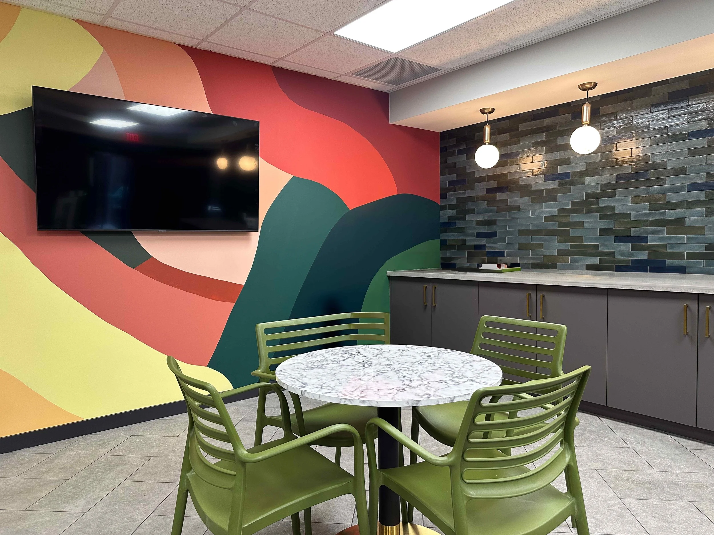 Corporate break room with bold colorful abstract mural wall in red, orange, yellow and teal, mounted TV, zellige tile kitchen wall with brass globe pendants, gray cabinetry, and sage green café chairs, interior design by Luxe Style Studio