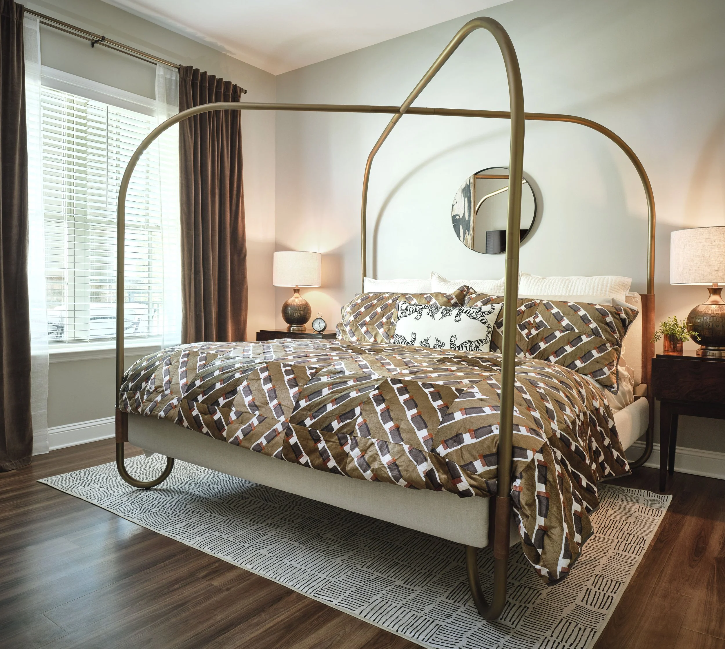 Front view of the West Broad primary bedroom with an arched brass canopy bed, layered geometric and animal-print bedding, brown velvet drapery, dark dresser, and ceramic lamps, designed by Ashley Schneider