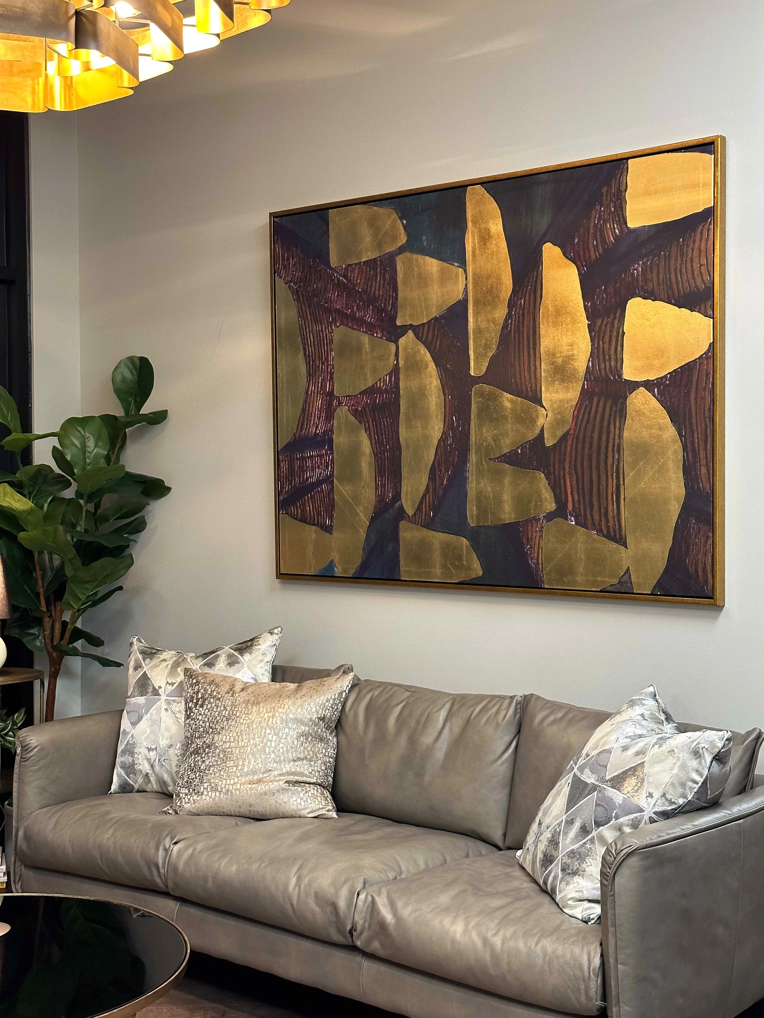 Corporate office lounge seating area with taupe leather sofa, metallic accent pillows, large gold leaf abstract artwork in brass frame, fiddle leaf fig tree, and brass multi-light chandelier above, interior design by Luxe Style Studio