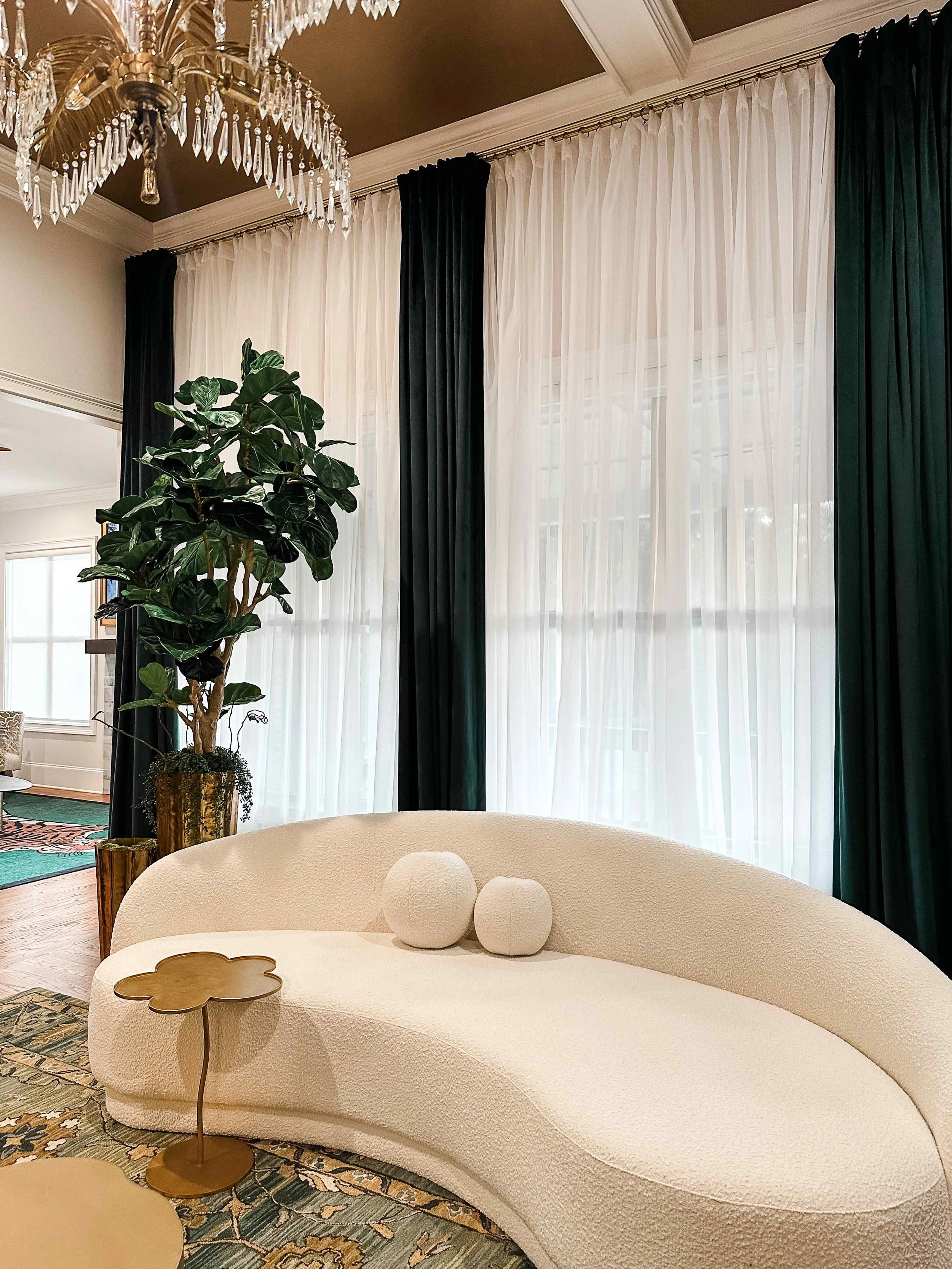 Noble living room in East Cobb with a curved cream bouclé sofa, layered emerald velvet and sheer drapery, and crystal chandeliers beneath a dark coffered ceiling, designed by Luxe Style Studio