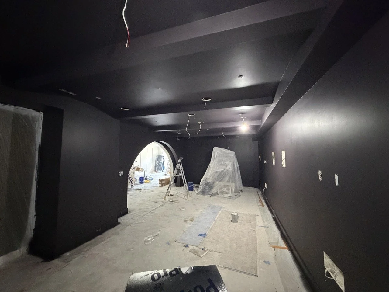 Home theater renovation in progress with dark painted walls, arched opening, protected floors, and wiring ready for lighting fixtures