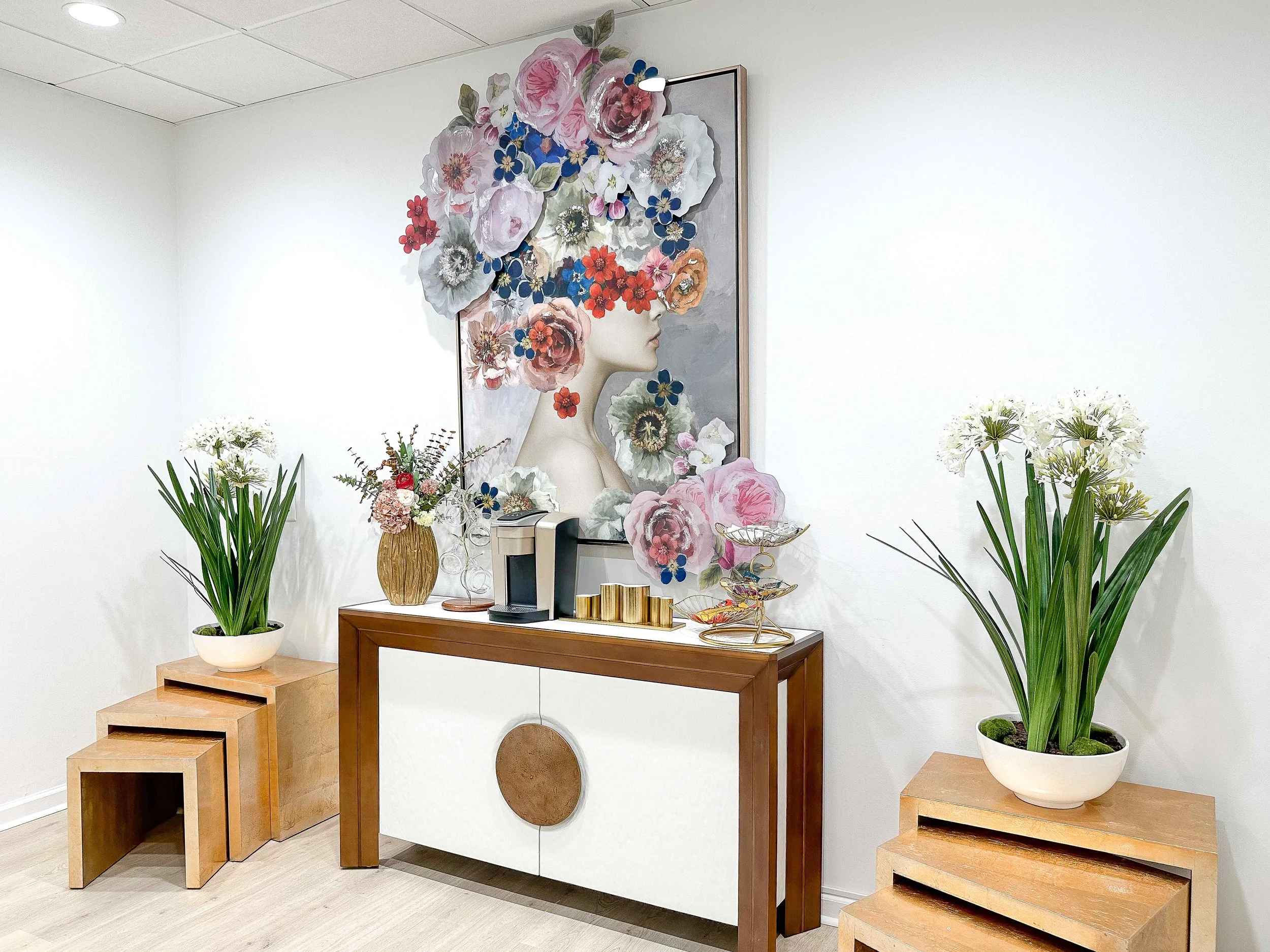 Medical spa reception console styled with gold vase, coffee station, and brass accessories beneath oversized dimensional floral portrait art, flanked by natural wood nesting tables and lush botanicals, interior design by Luxe Style Studio