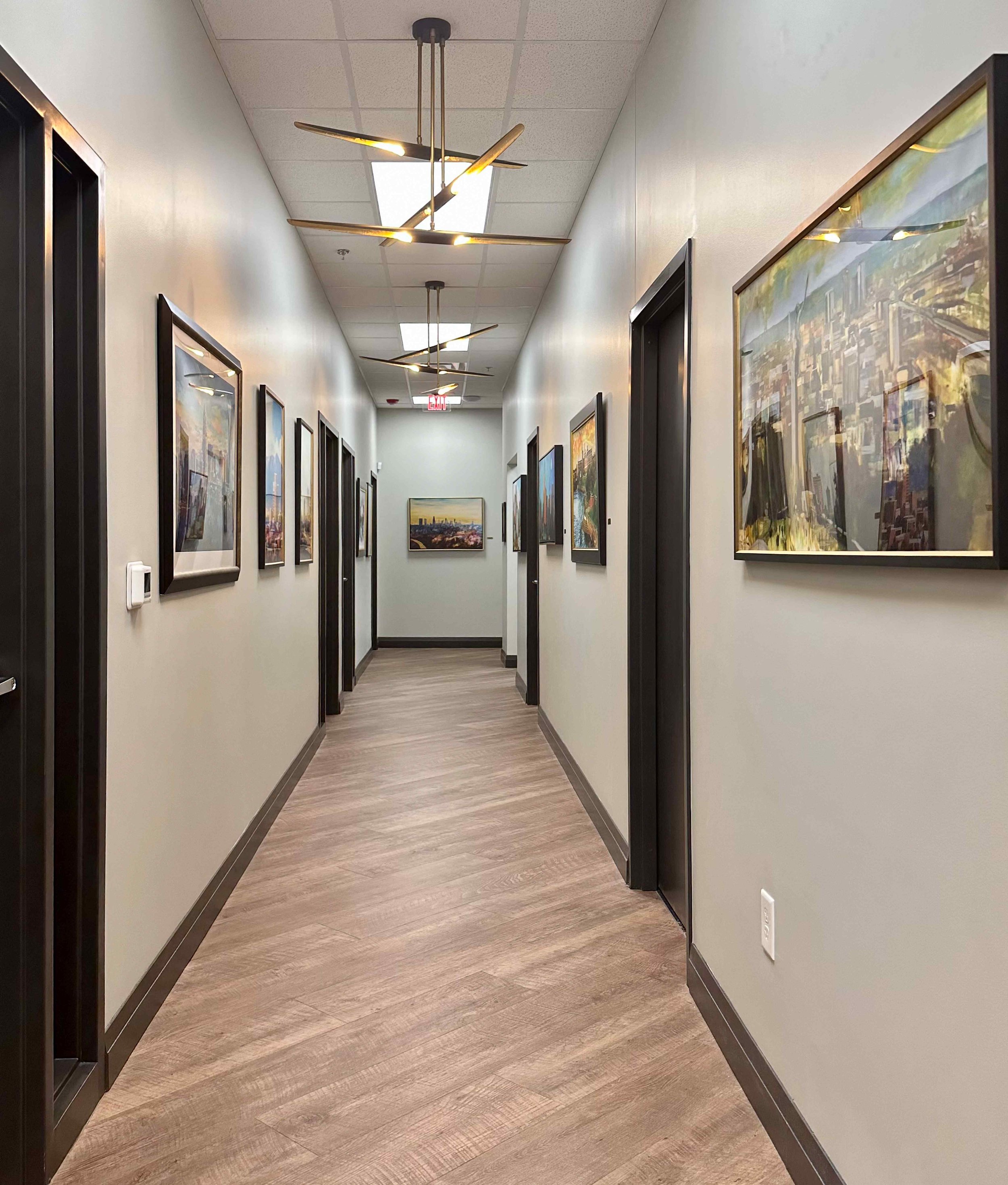 Corporate office hallway styled as an art gallery with large-scale cityscape paintings in black frames, brass sputnik pendant lights, wood plank floors, and dark framed office doors, interior design by Luxe Style Studio