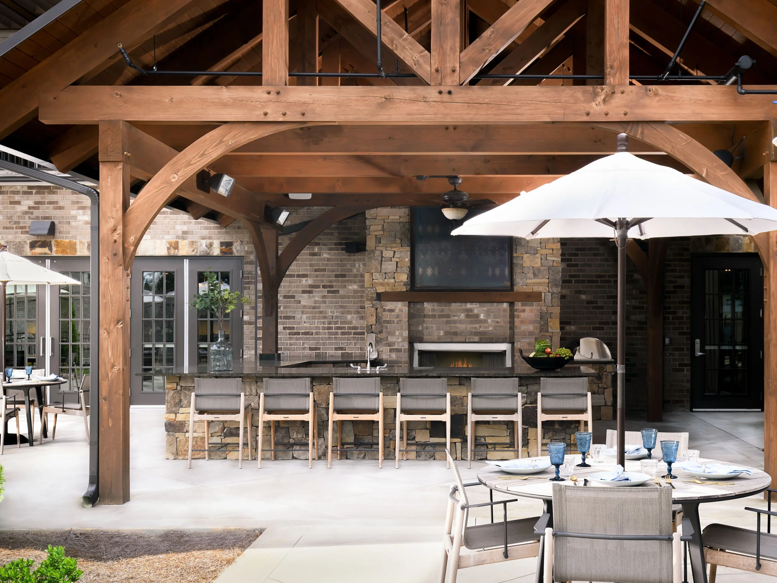 Local Hearth outdoor bar in Acworth with a stacked-stone base, dark counter, teak stools, timber pergola, outdoor fireplace, and umbrella-shaded dining tables, designed by Ashley Schneider