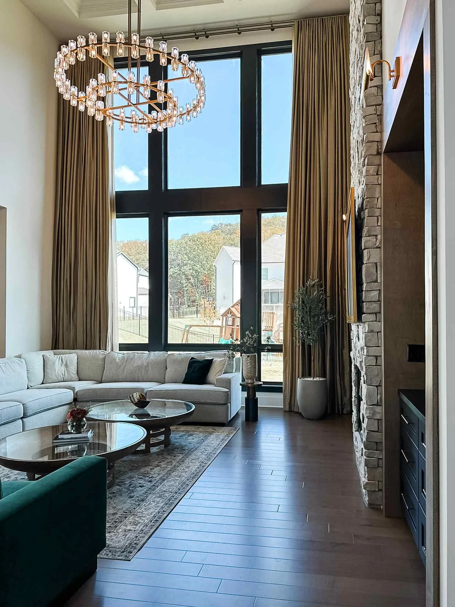 Two-story great room with oversized crystal ring chandelier, olive velvet drapes, and stone accent wall by Luxe Style Studio